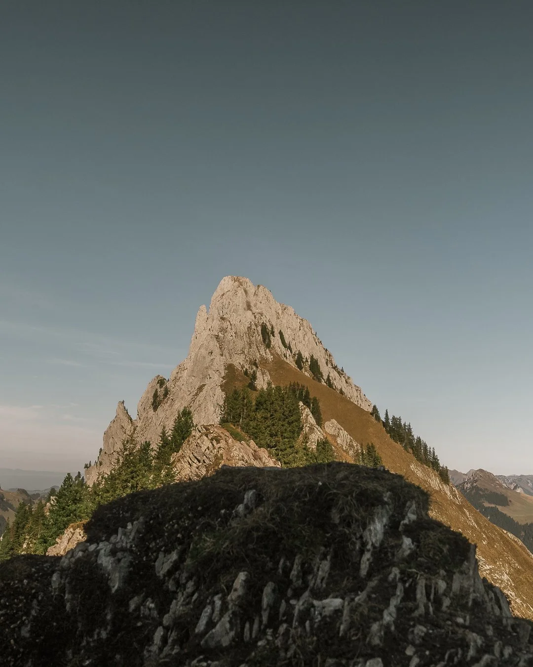 A mountain with a pointed peak, surrounded by trees and a rocky foreground under a clear sky during daytime.