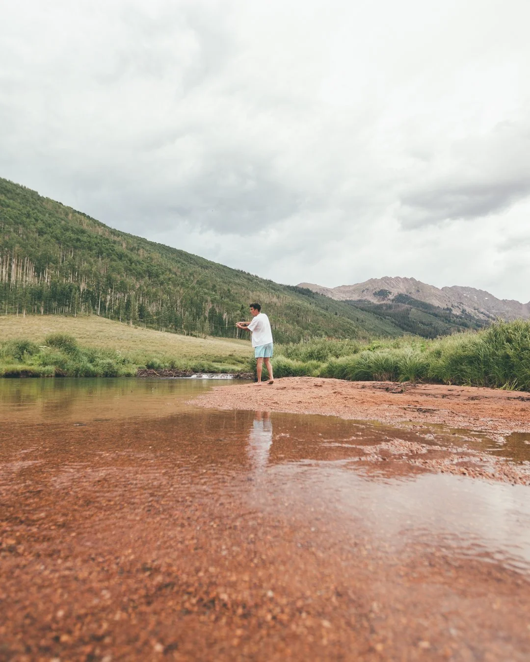 Person standing in a shallow river, holding a fishing rod, with green mountains and cloudy sky in the background.
