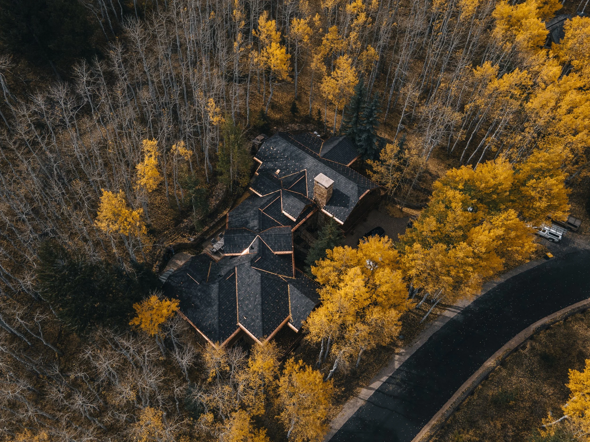 Aerial view of a house surrounded by trees with fall foliage, some trees without leaves, and a parking lot with cars nearby.