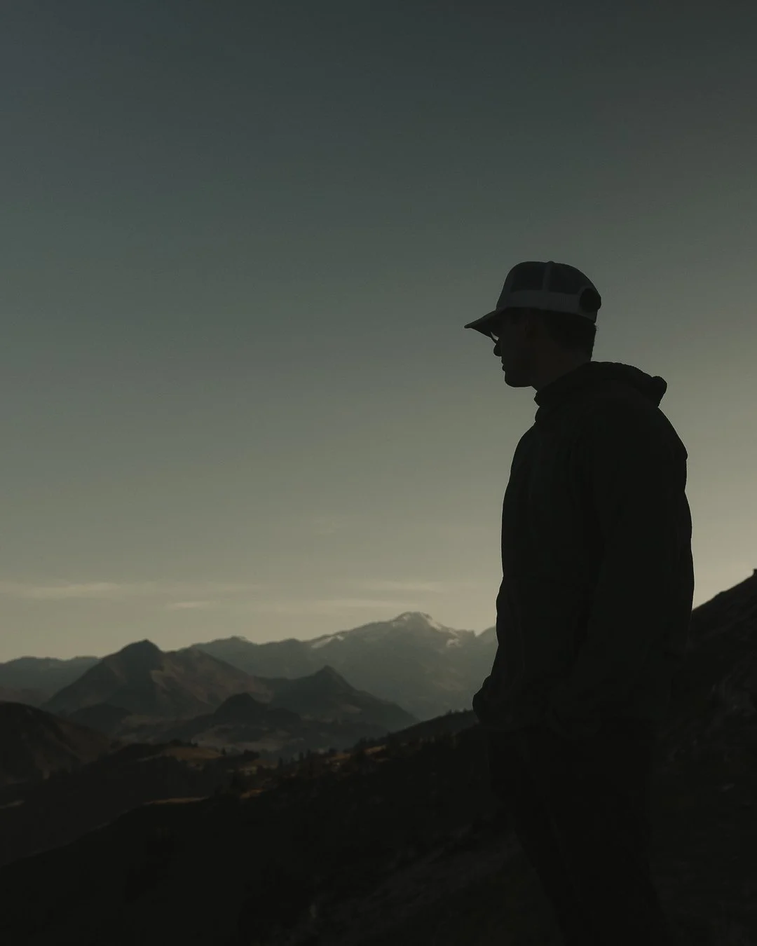 Silhouette of a person wearing a cap and hoodie, standing on a mountain with a mountain range and snow-capped peaks in the background during dusk or dawn.