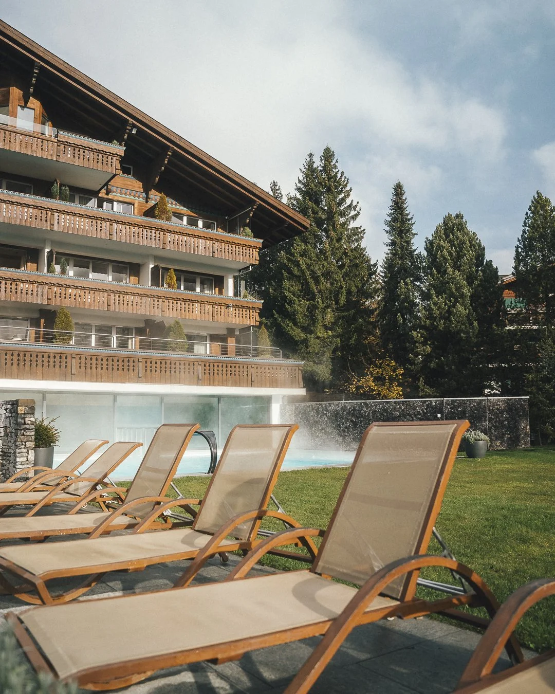 Ermitage Wellness & Spa Hotel in Gstaad, Switzerland. Sun loungers next to a swimming pool in front of a multi-story wooden building with balconies, surrounded by tall trees.
