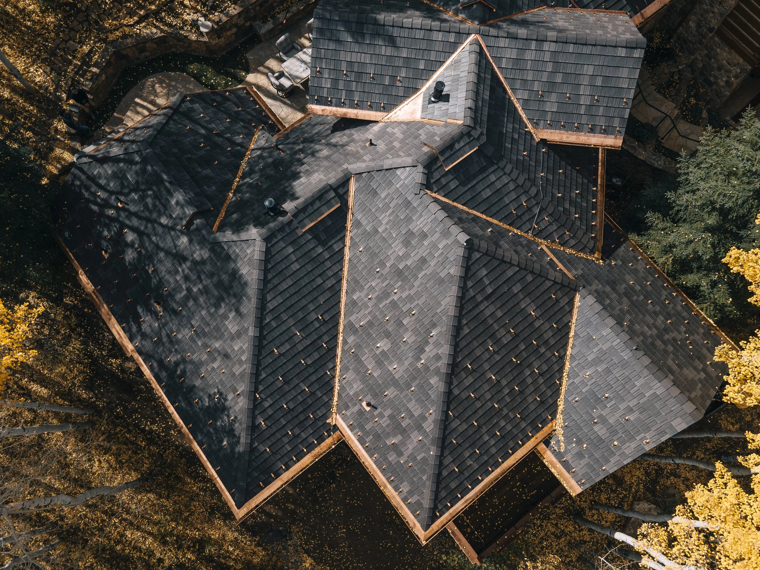 An aerial view of a house with a gray shingled roof, surrounded by trees with yellow leaves and a backyard with outdoor furniture.