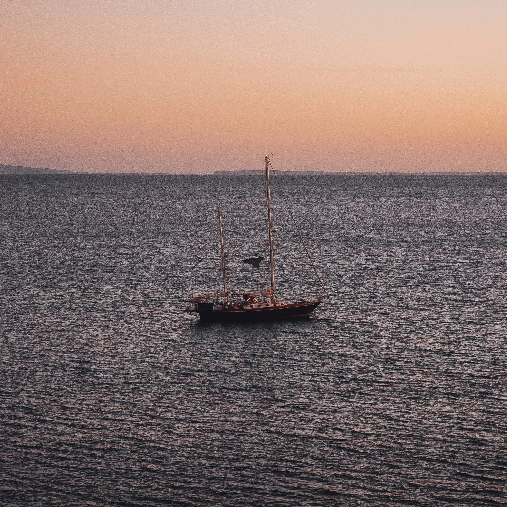 A sailboat on calm sea waters during sunset with a pinkish sky and a distant landmass on the horizon, shot in Ibiza