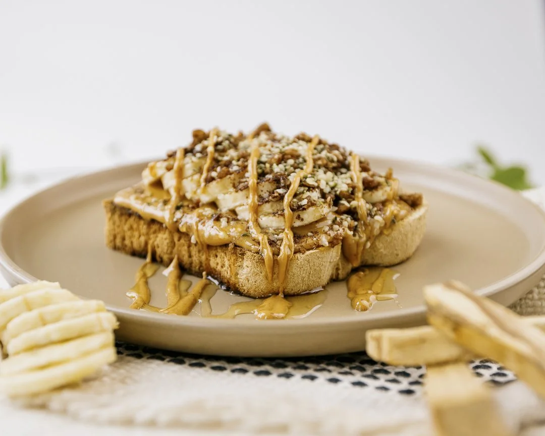 A slice of bread topped with banana slices, peanut butter, chocolate, chopped nuts, and caramel sauce, served on a beige platefor Innerlight Juice.