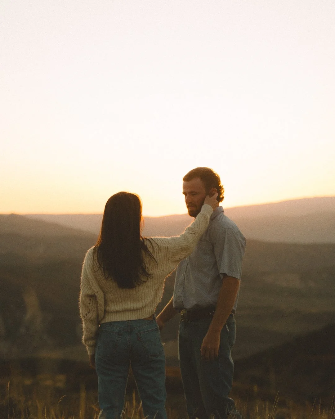 A woman touches the face of a man outdoors during sunset, with rolling hills in the background.