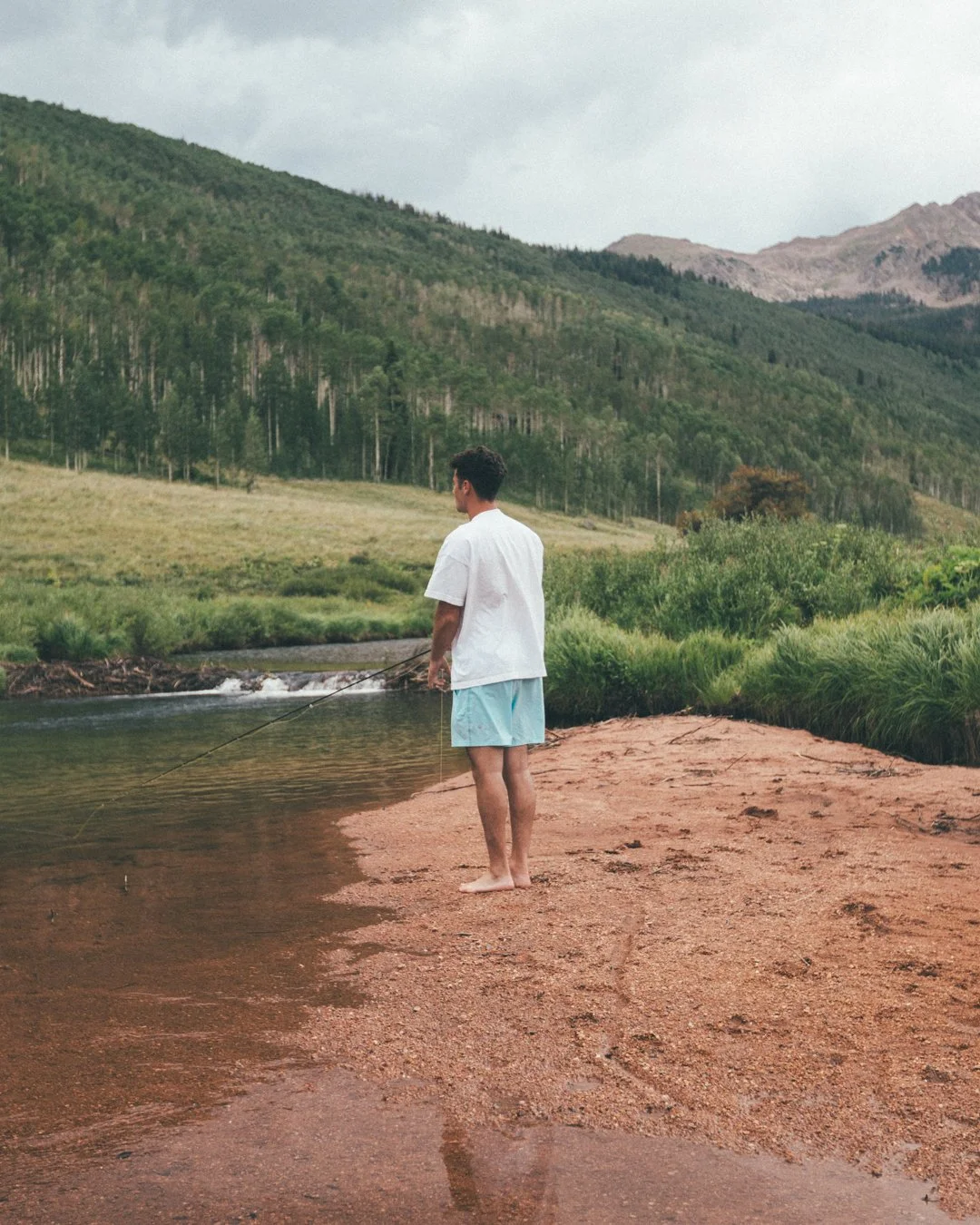A person standing barefoot on a sandy riverbank, fishing in a peaceful river with green hills and mountains in the background.