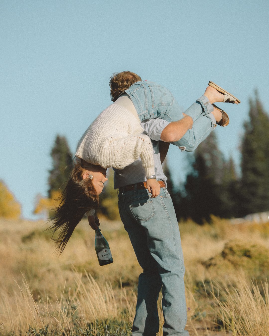 A person giving a piggyback ride to a woman in a field with trees in the background on a sunny day.