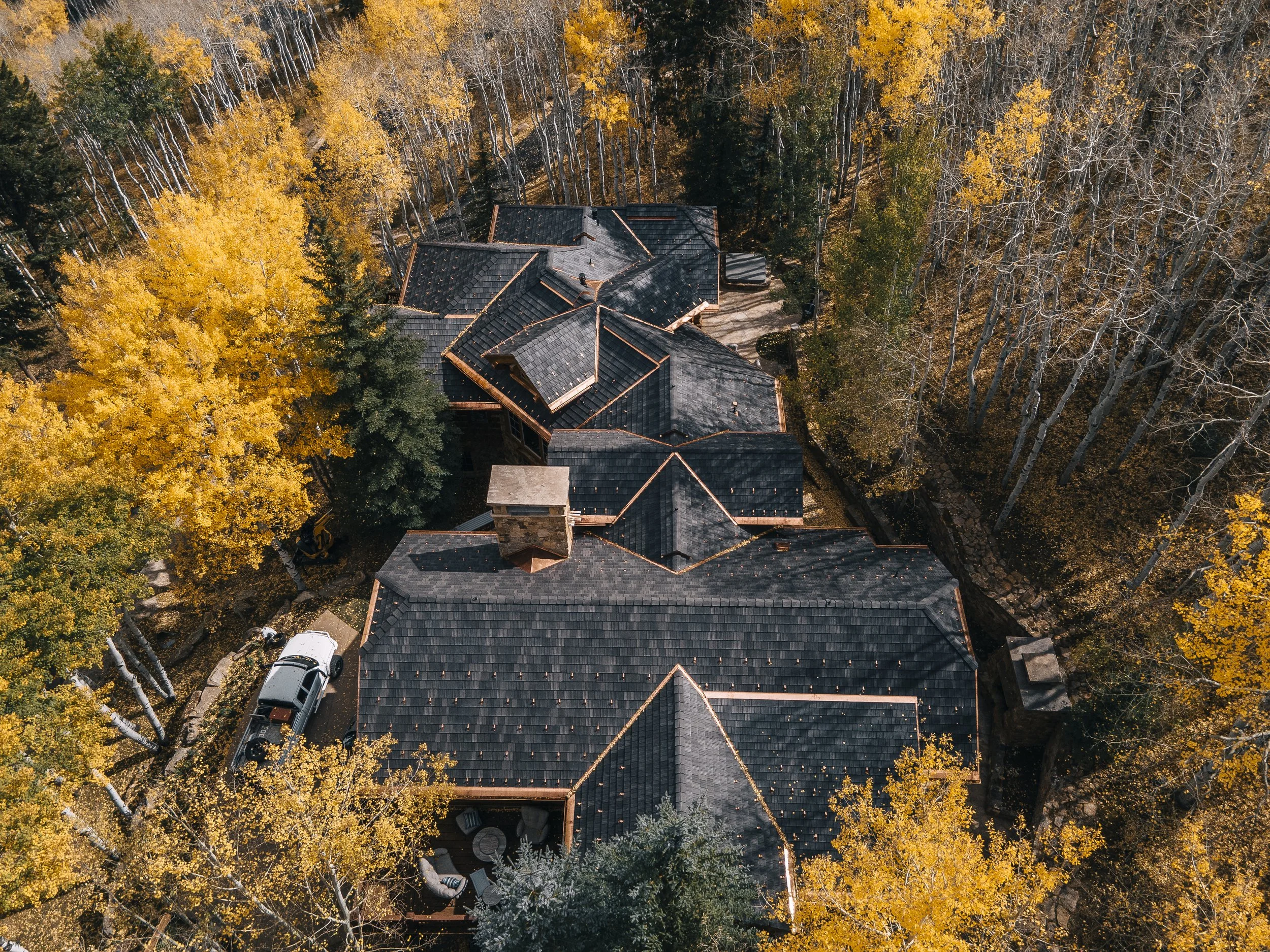 An aerial view of a house surrounded by trees with autumn-colored leaves, some with bare branches, and a driveway with parked cars.