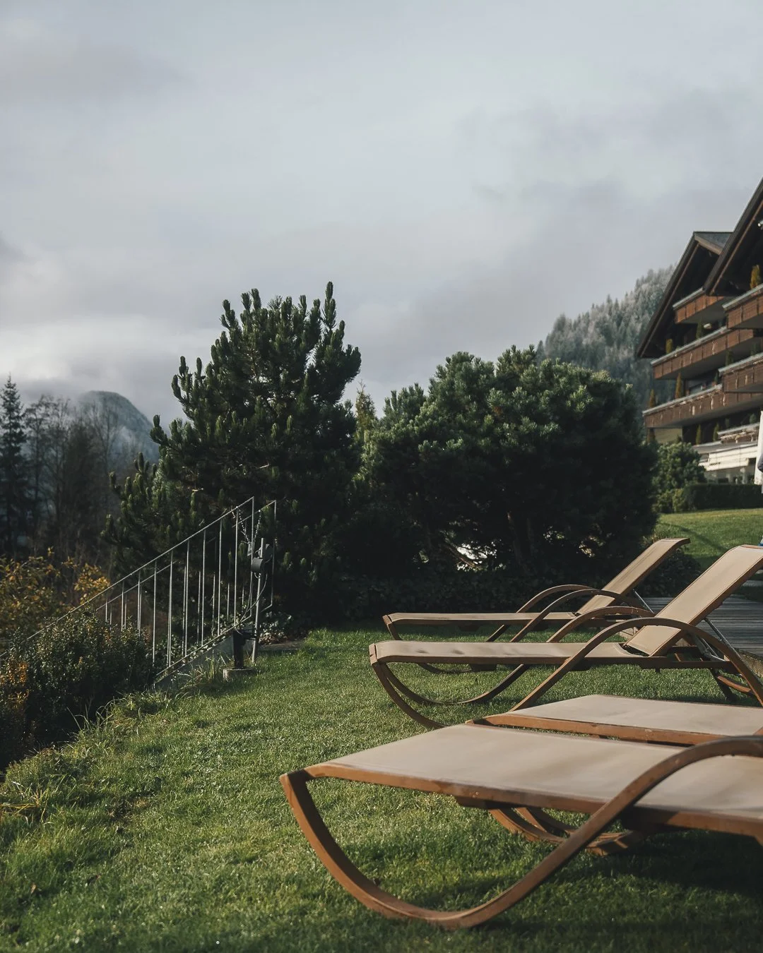 Three wooden lounge chairs on a grassy area next to a swimming pool, with trees, shrubs, and a multi-story building in the background on a cloudy day.