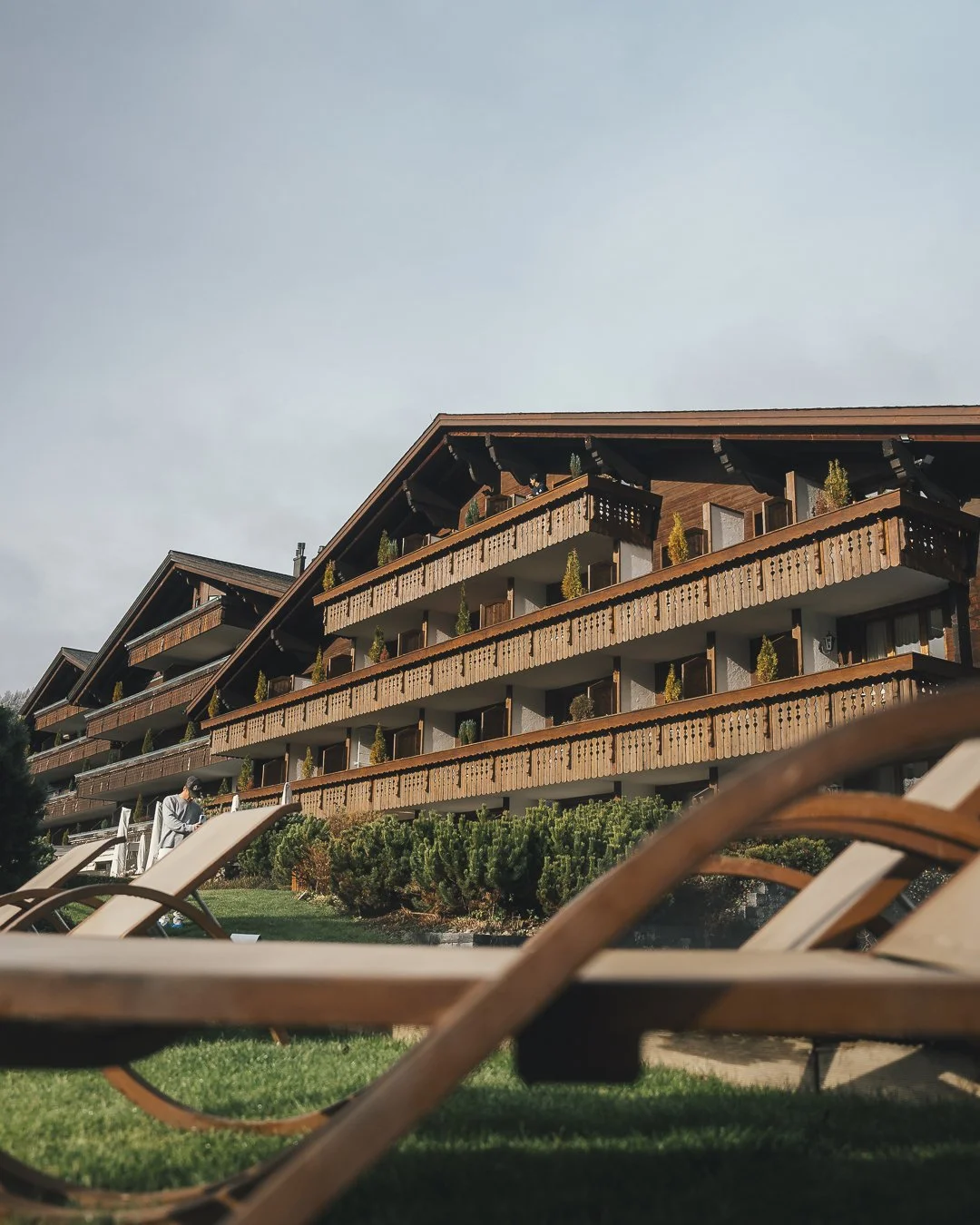 Multi-story wooden chalet-style hotel with balconies, lush landscaping, and outdoor lounge chairs in the foreground.