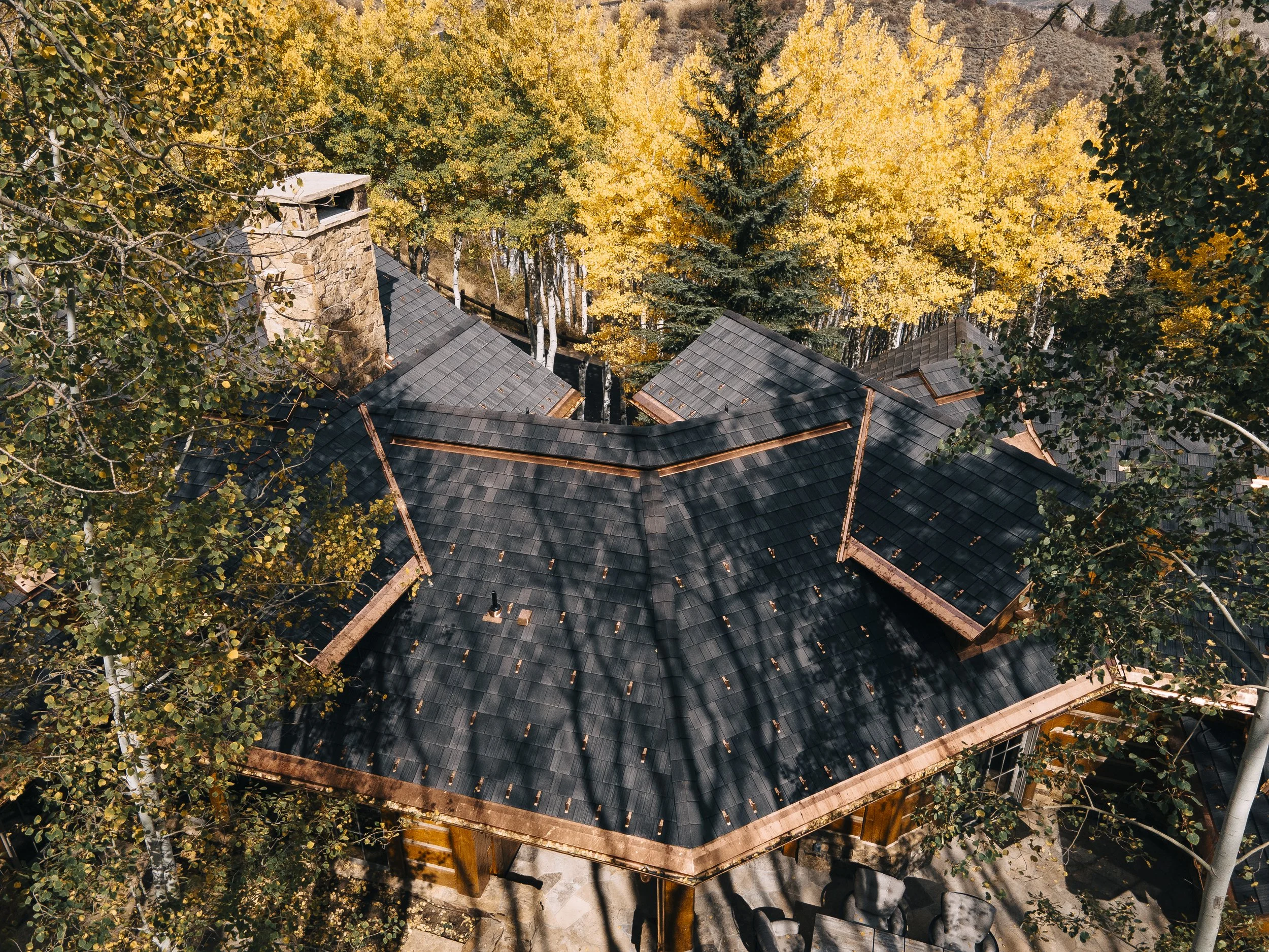 A bird's eye view of a house roof surrounded by yellow autumn trees. The roof has a chimney and multiple sections covered with dark shingles.