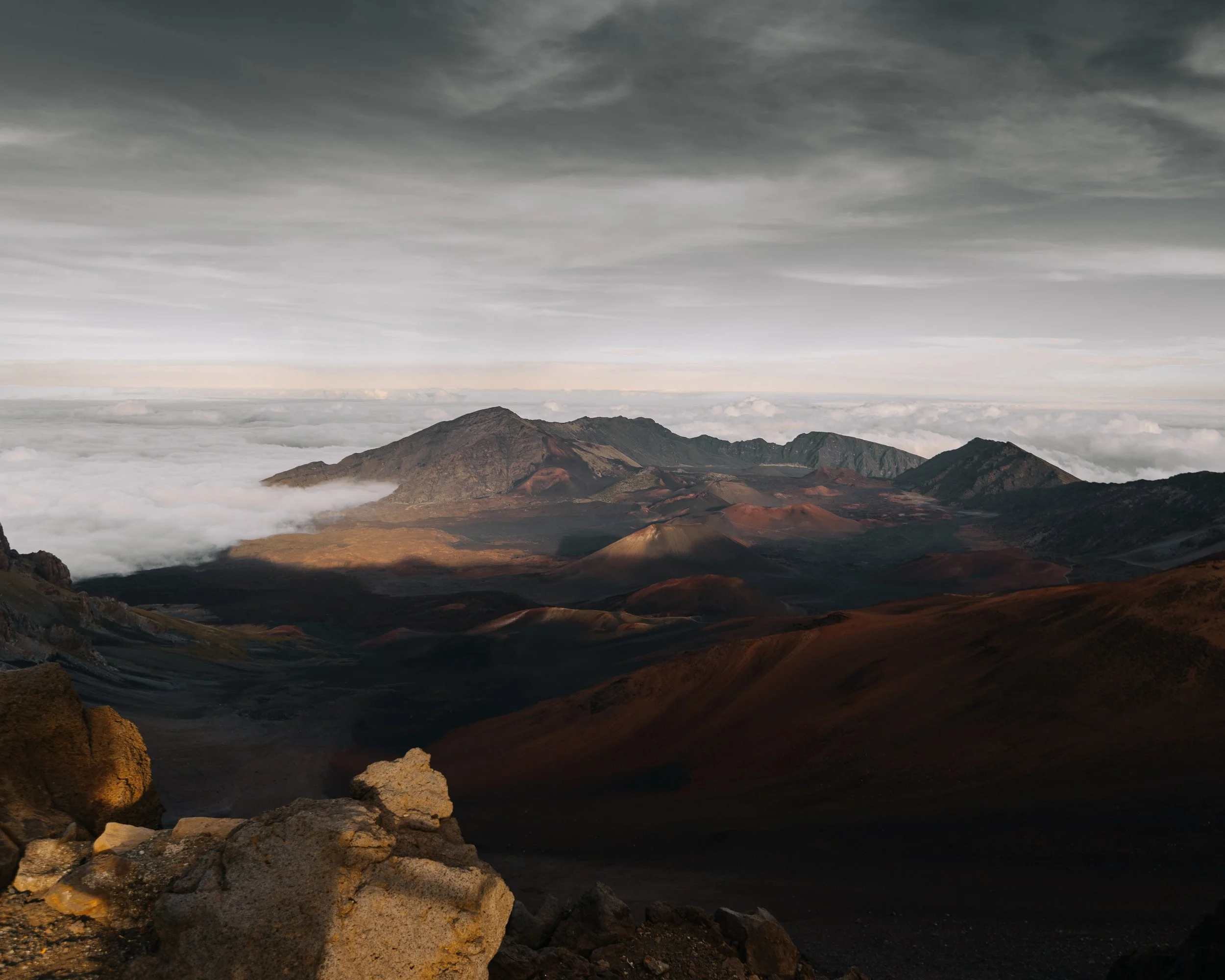 View of a mountain landscape with clouds below and dark rugged mountain peaks in the foreground under a cloudy sky.