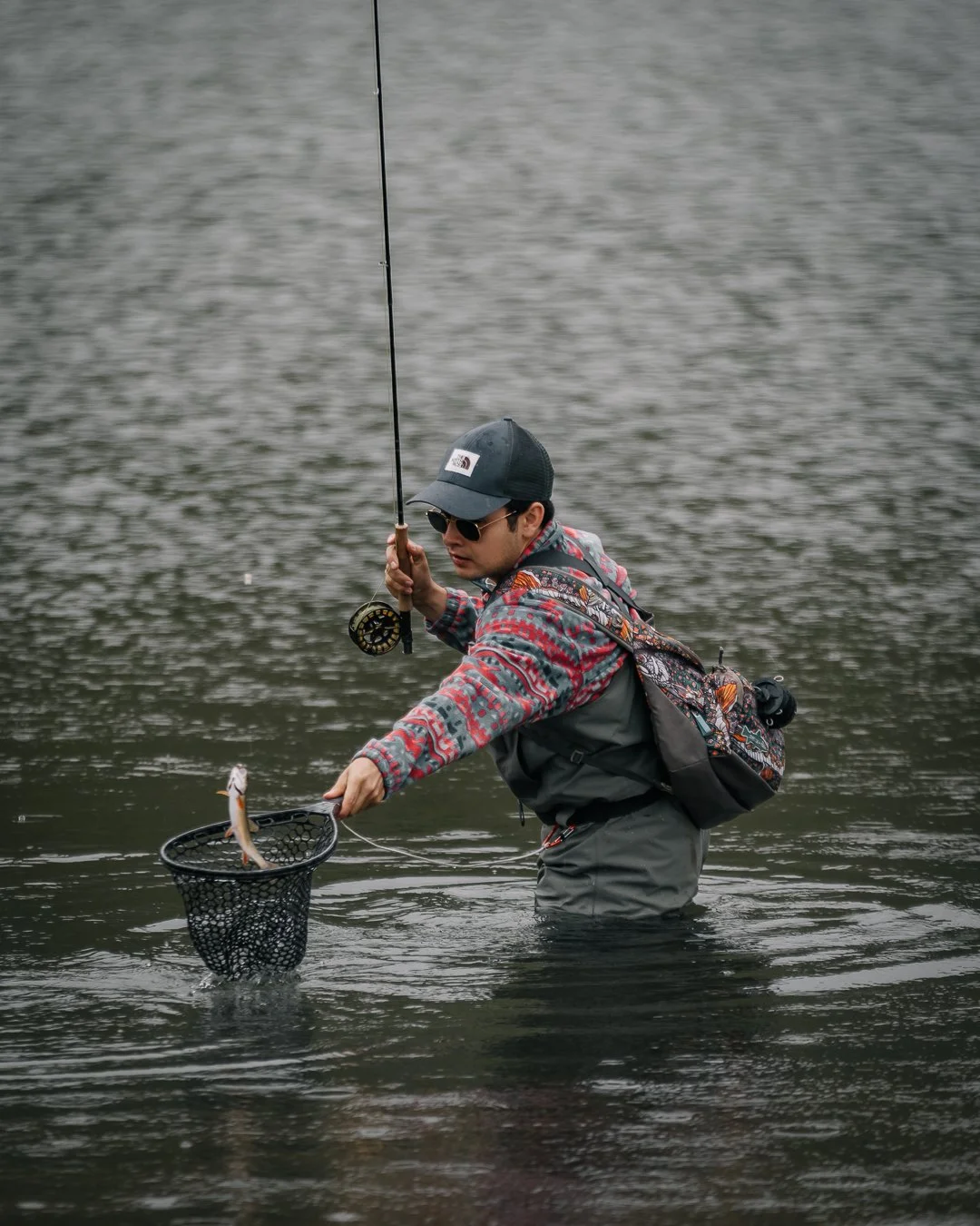 A person fishing in a river, holding a fishing rod in one hand and a net with a small fish in the other.