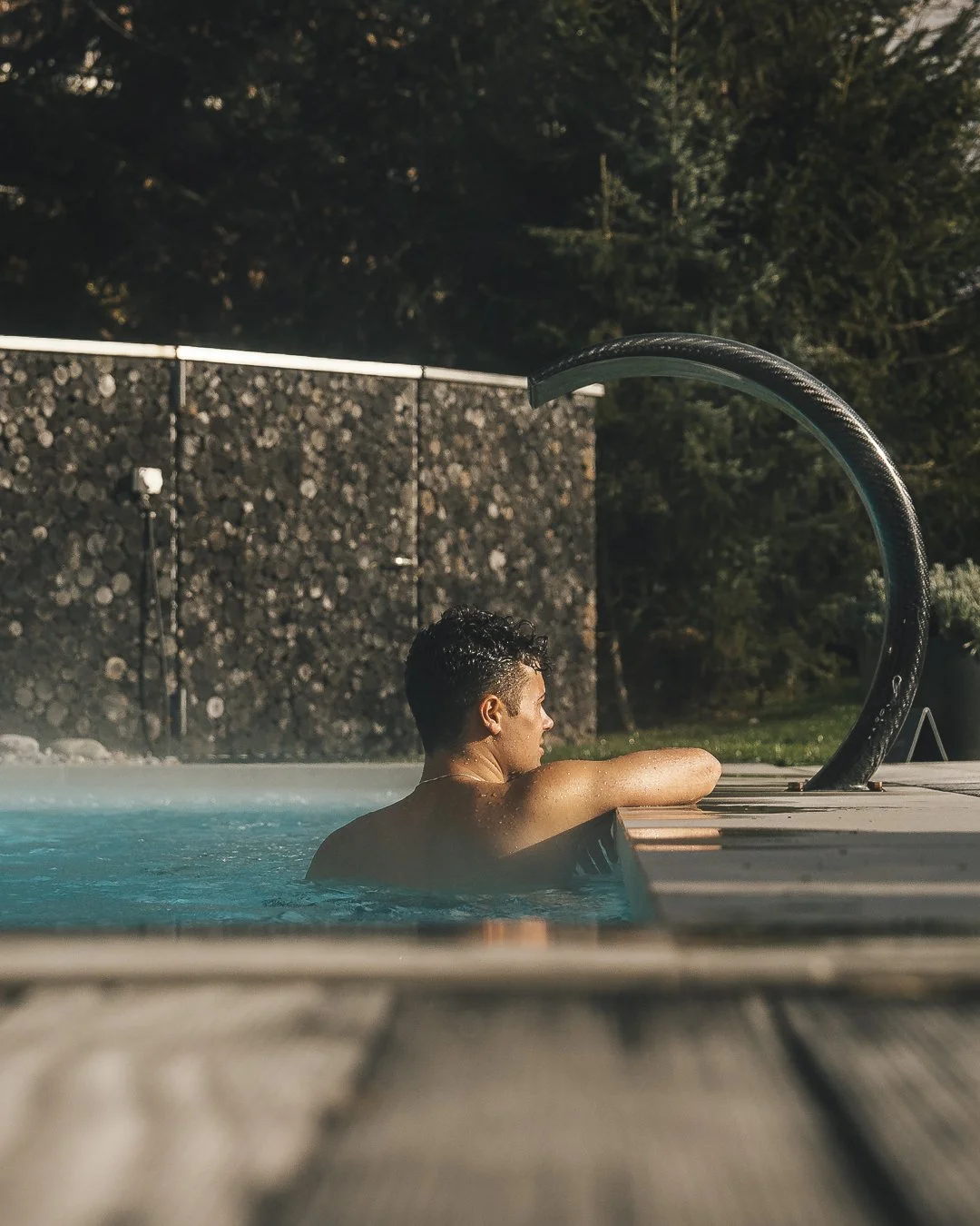 Ermitage Wellness & Spa Hotel in Gstaad, Switzerland. A man relaxing in an outdoor swimming pool during the evening, leaning on the pool edge with trees and a log-covered fence in the background.