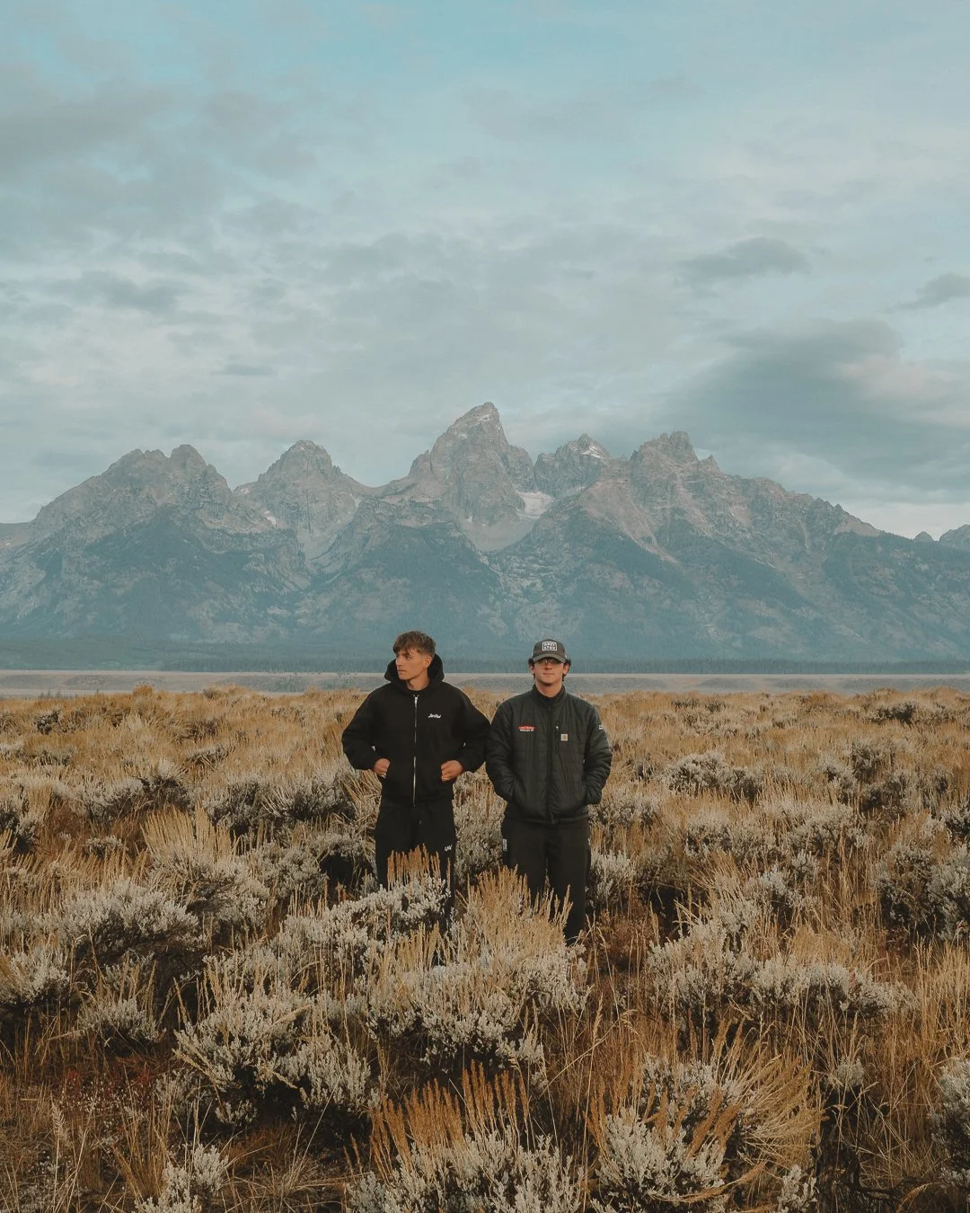 Two men standing in a desert landscape with mountains in the background, wearing black jackets.