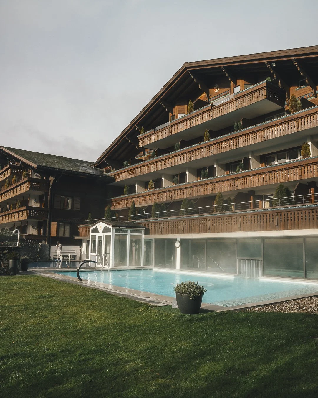 Ermitage Wellness & Spa Hotel in Gstaad, Switzerland. A multi-story wooden hotel with balconies and a rooftop garden, overlooking an outdoor swimming pool with steam rising from it, surrounded by a well-manicured lawn and potted plants.