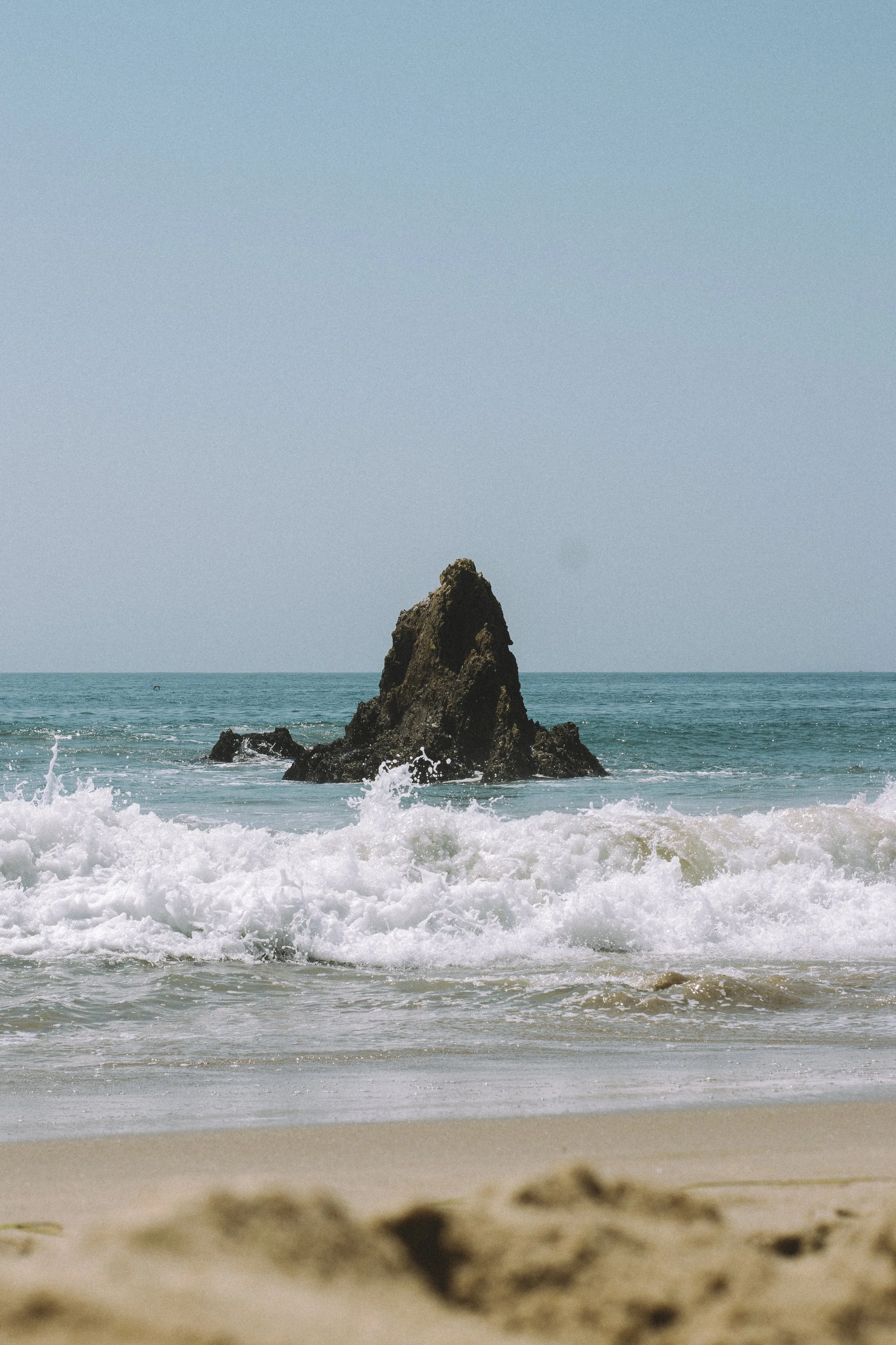 A large rock formation rising from the ocean waves near the shoreline, with a clear blue sky overhead.