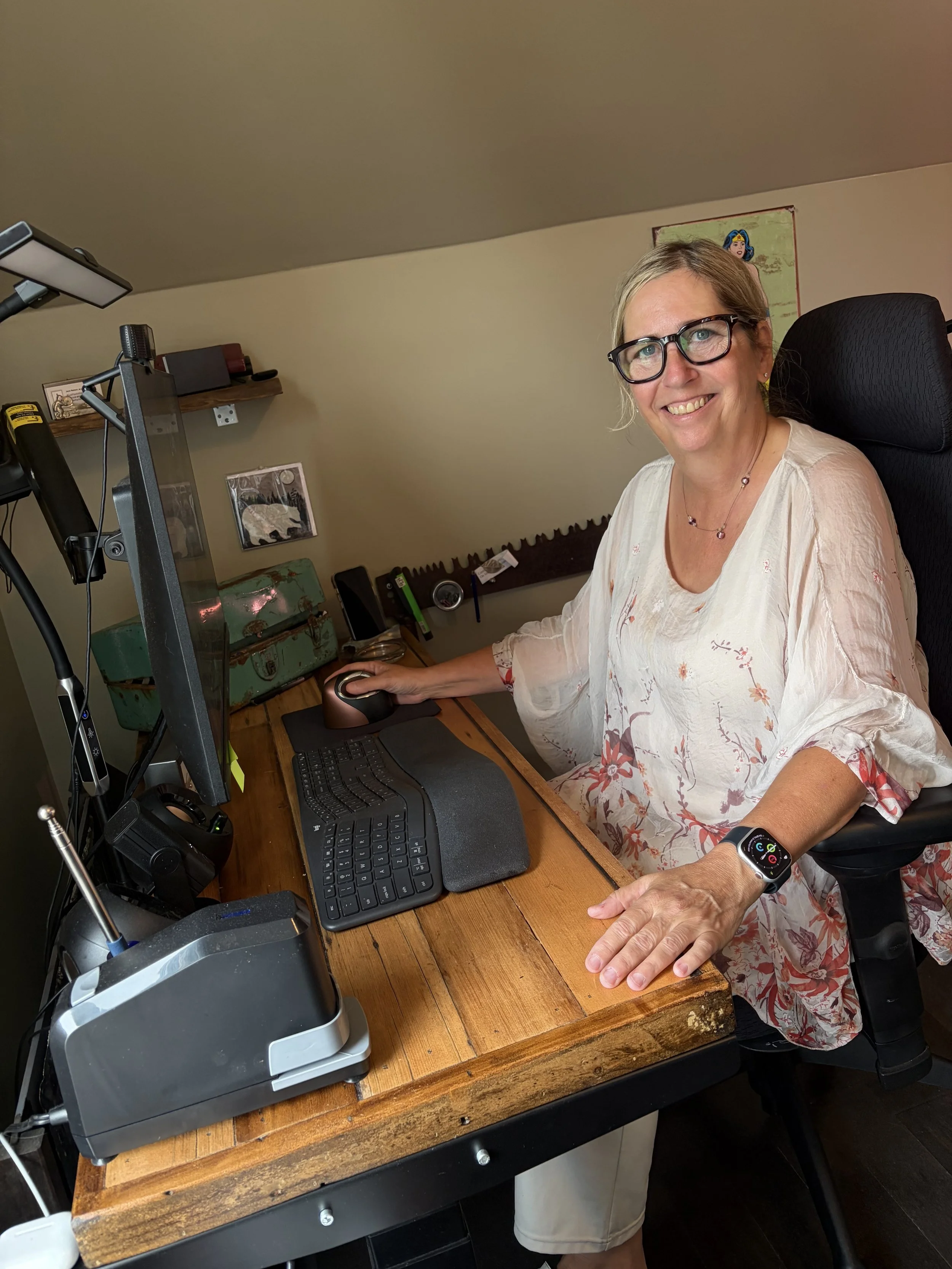 A woman with glasses and a floral blouse sitting at a wooden desk, smiling, using a computer and wearing a smartwatch.