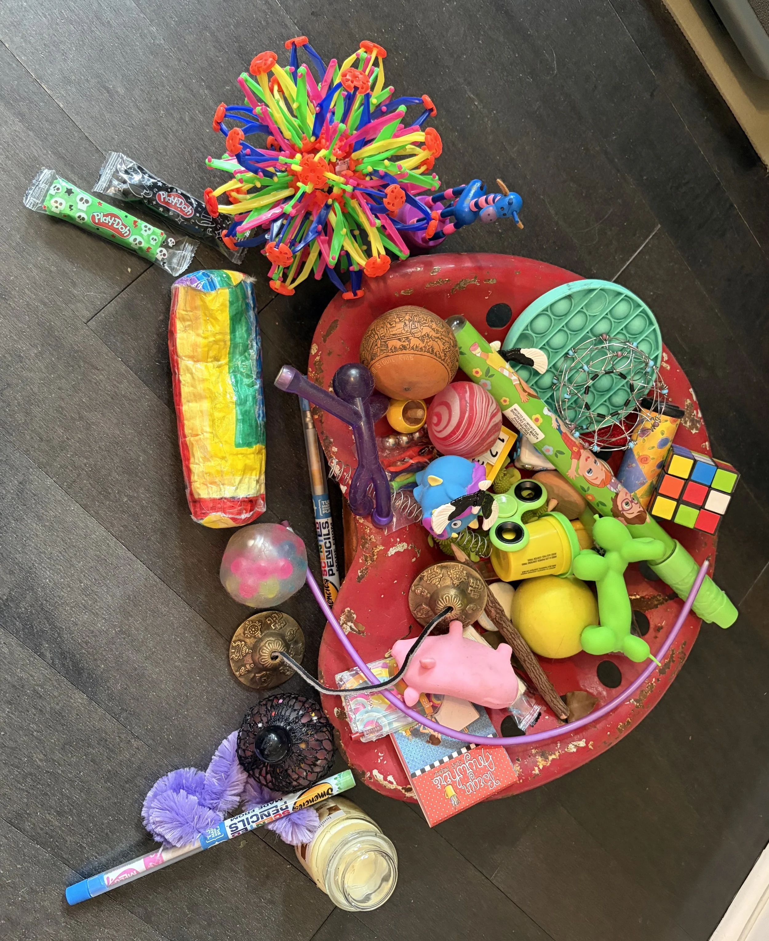 Cluttered table with various toys, including a rainbow-colored slap bracelet, a rubber chicken, maracas, a Rubik's Cube, a blue and pink toy horse, a plastic ball, a mesh ball, and a plastic container with white substance, all on a dark wooden floor.
