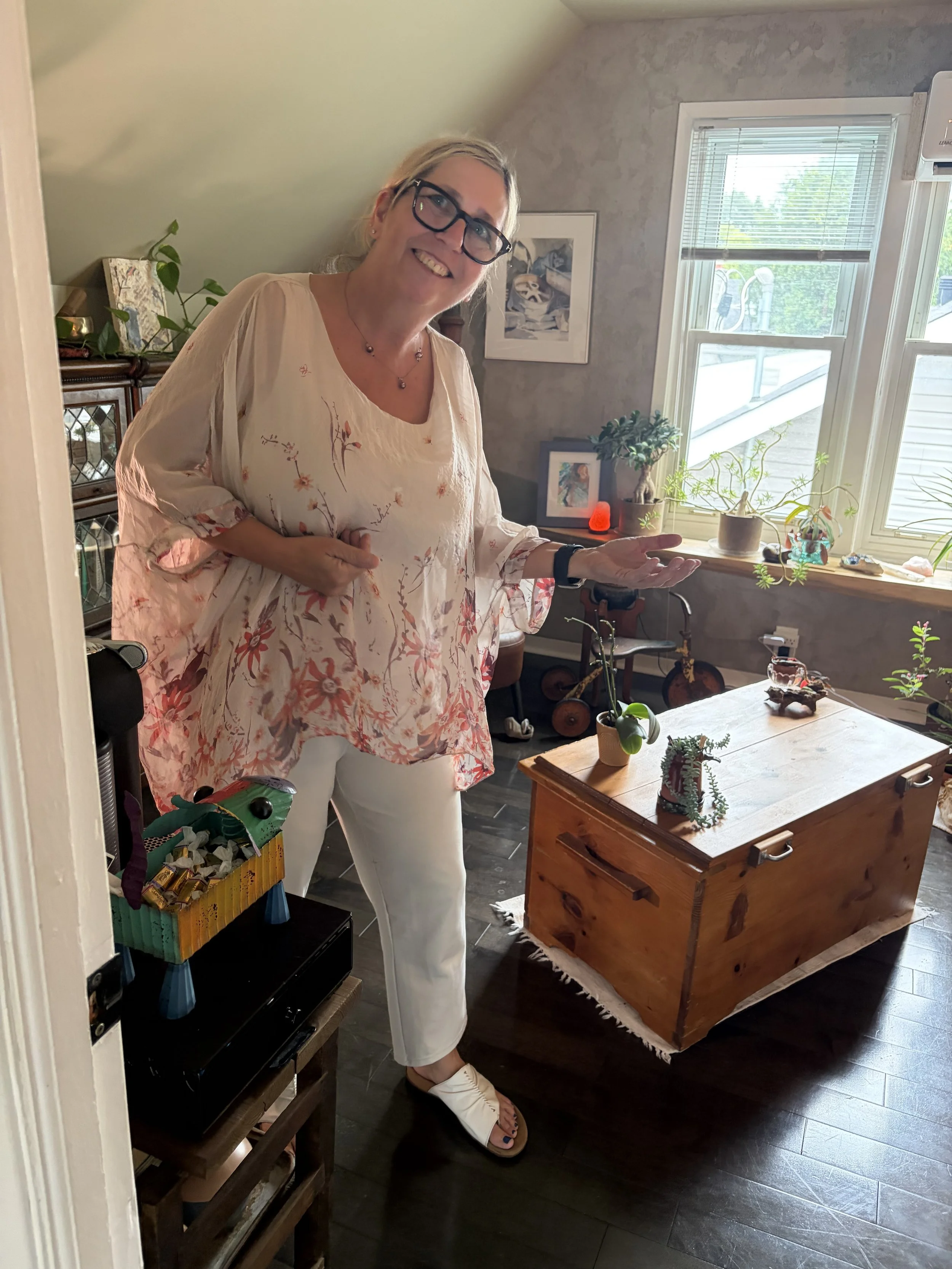 A woman with blonde hair, glasses, wearing a floral blouse and white pants, standing in a room with various houseplants and decorations, smiling, and gesturing with her right hand toward a window. The room has gray walls, a wooden chest, framed pictures, and a window with blinds letting in natural light.