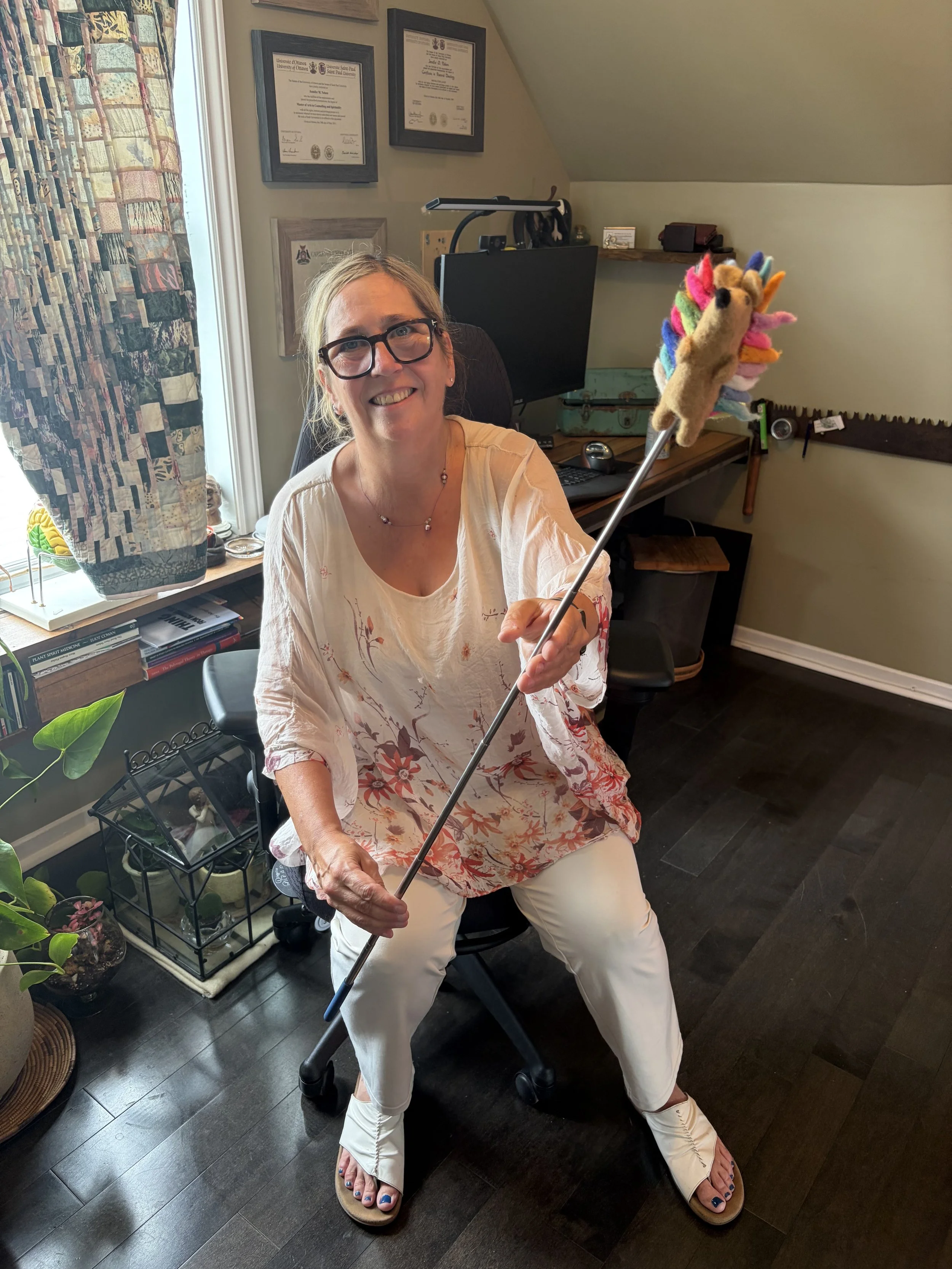 A smiling woman with glasses and blonde hair, wearing a floral blouse and white pants, sitting on a walker and holding a stick with colorful felted animals attached, in a home office with framed diplomas on the wall, a desk with a computer monitor, and potted plants.
