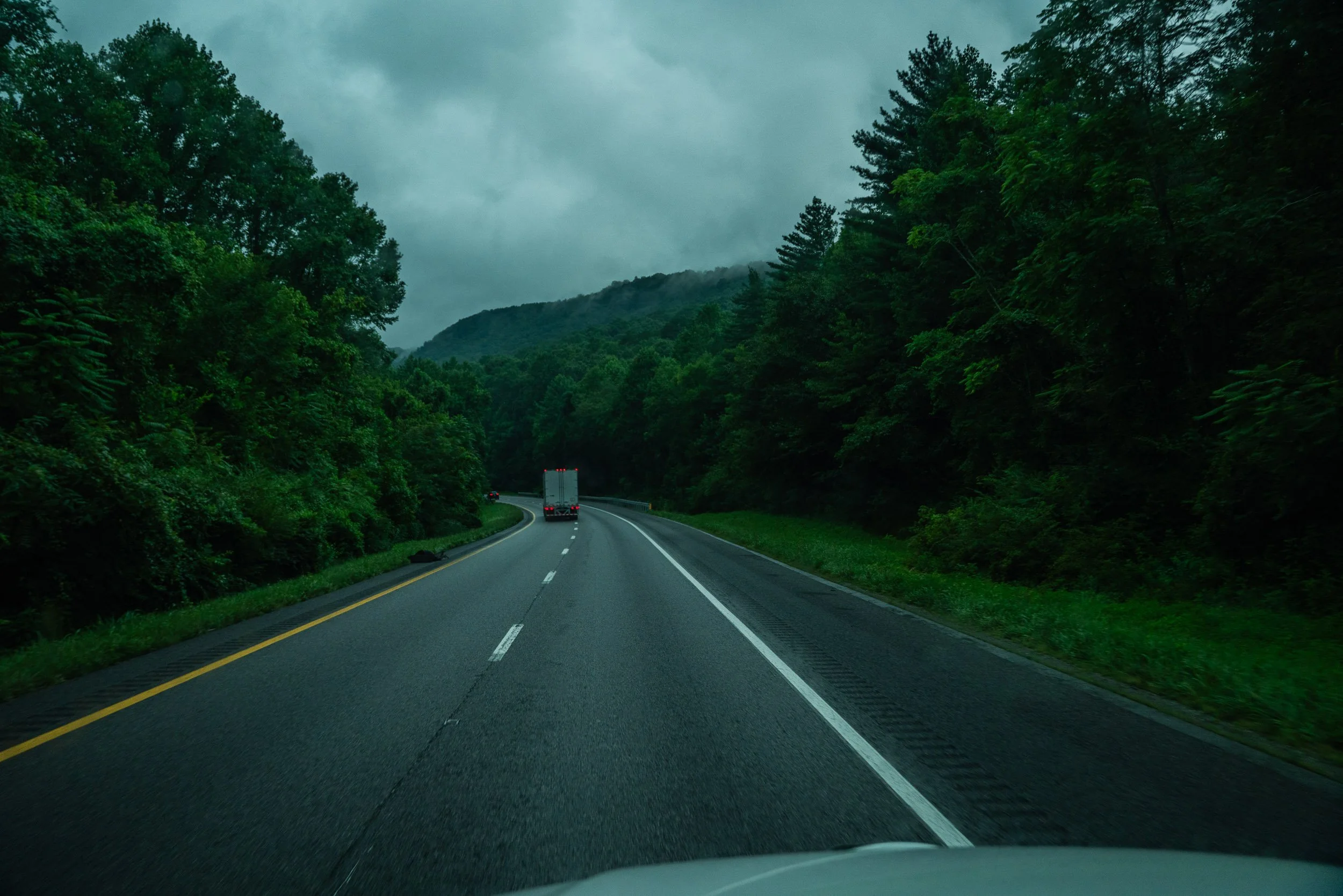 A scenic view of a winding highway surrounded by dense green trees on both sides, with hills or mountains in the background and cloudy skies overhead.