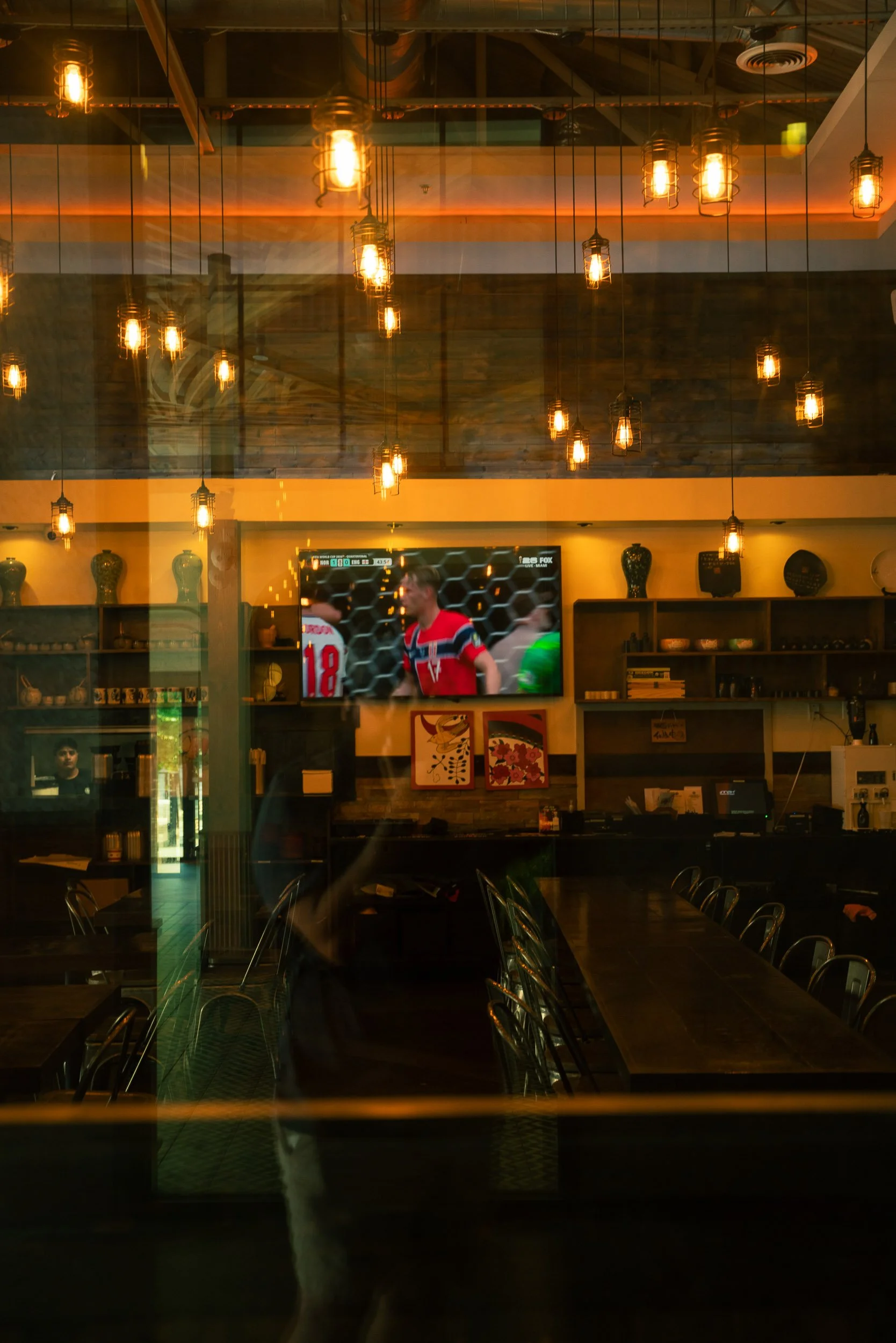 Interior of a restaurant or bar with exposed hanging filament light bulbs, a large television screen showing a sports game, and wooden shelves with vases and decor. The photo is taken through a glass window or door, reflecting some of the interior.