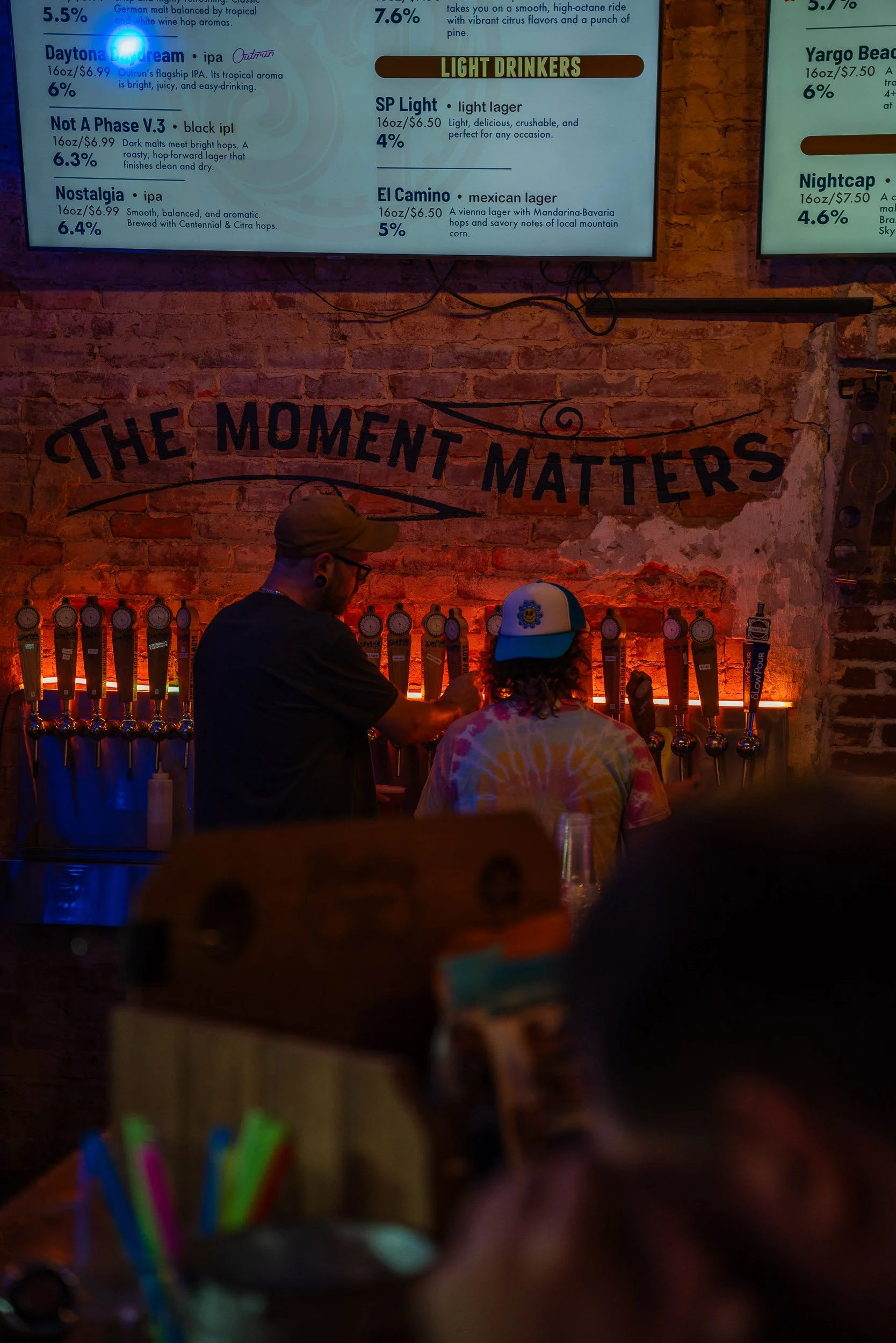 Bar area at a restaurant or bar with a sign on the brick wall reading 'The Moment Matters,' two bartenders working, and a row of beer taps behind the counter. A man wearing a cap is serving a customer with curly hair and a tie-dye shirt. A document o