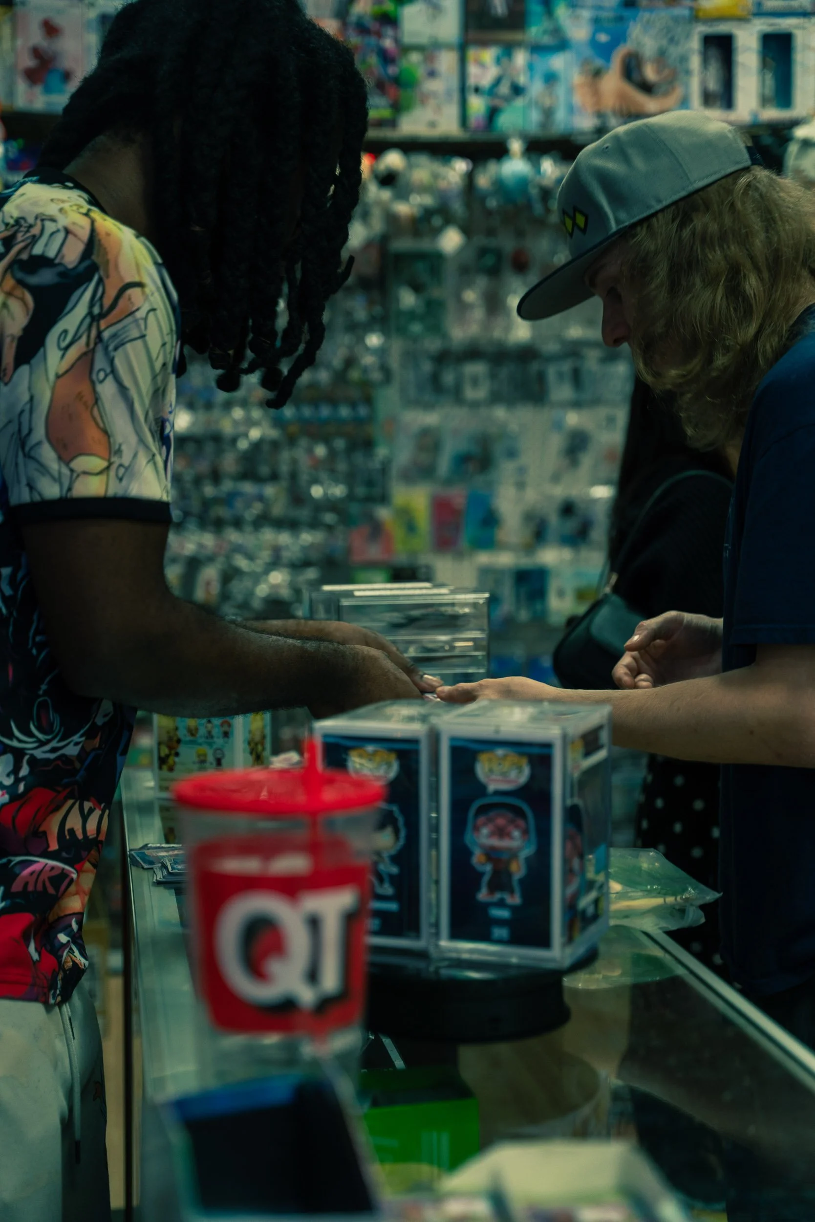 Two men, one with dreadlocks and colorful shirt, and the other with long hair, baseball cap, and jacket, are shopping at a store counter filled with various products, including Funko Pop figures, with shelves filled with toys and merchandise in the b