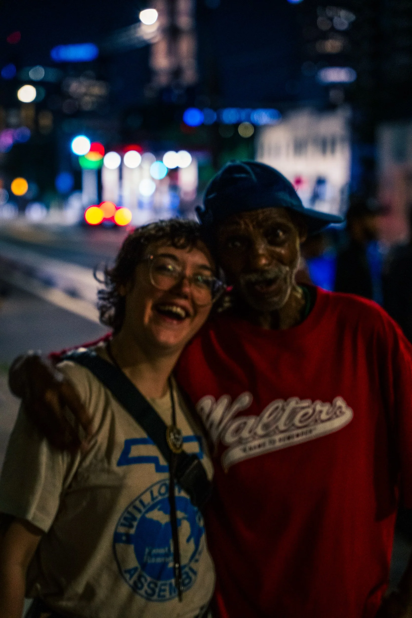 Two happy people, a woman with glasses and curly hair, and a man wearing a blue hat, smiling and standing close together at night on a city street with blurred colorful city lights in the background.