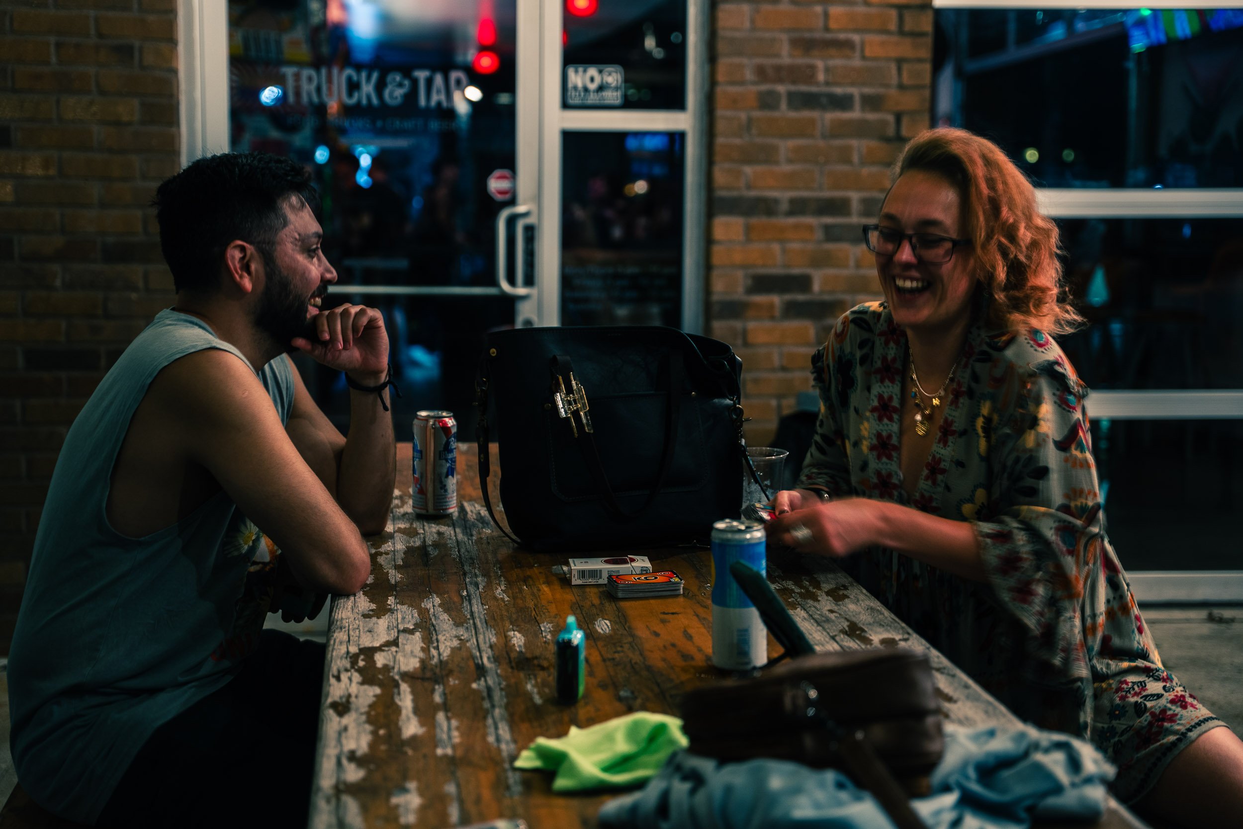 Two people, a man and a woman, sitting at a wooden table inside a restaurant or bar, laughing and enjoying each other's company. The man has a beard and short hair, wearing a sleeveless shirt, and the woman has curly hair, glasses, and is wearing a f