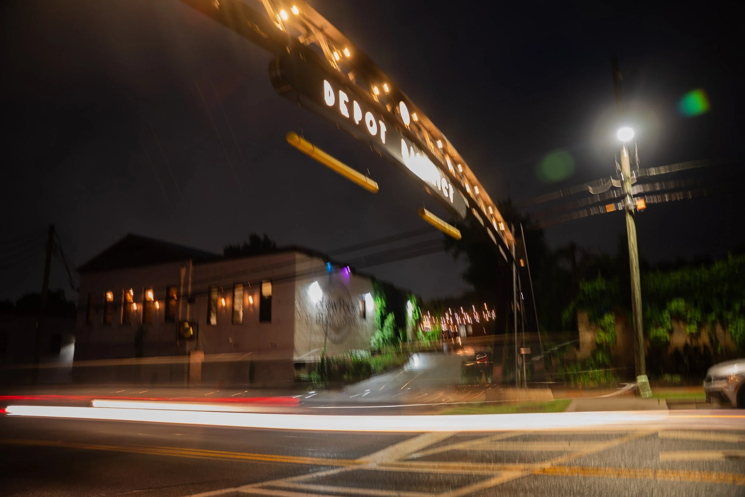 Nighttime scene with a blurred view of a street, showing streaks of light from moving vehicles, a street sign for Depot Street, and illuminated buildings in the background.