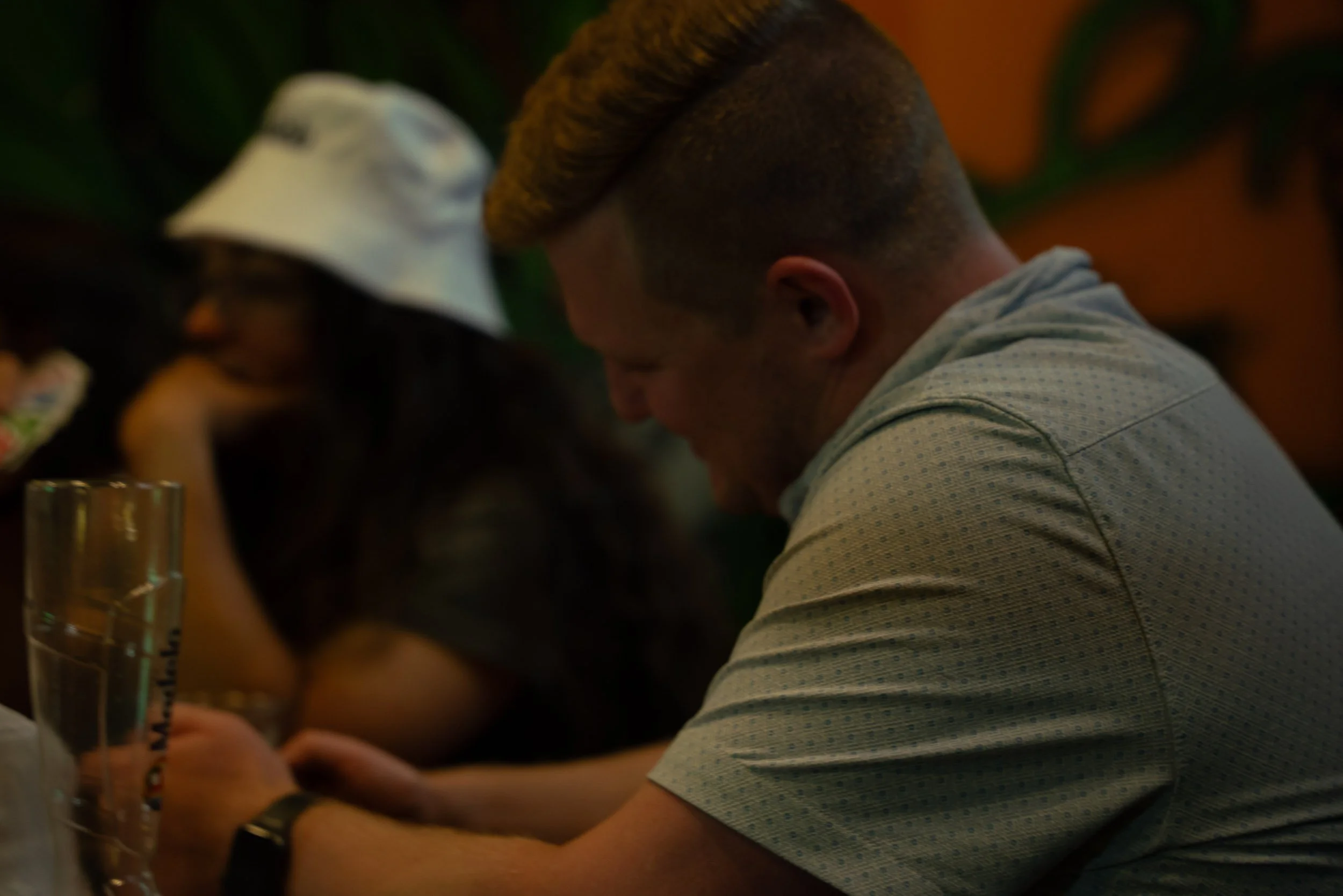 A person with short, curly blonde hair wearing a light grey t-shirt, sitting at a table in a dimly lit setting, with a woman wearing a white baseball cap and glasses in the foreground and a glass of water visible on the table.