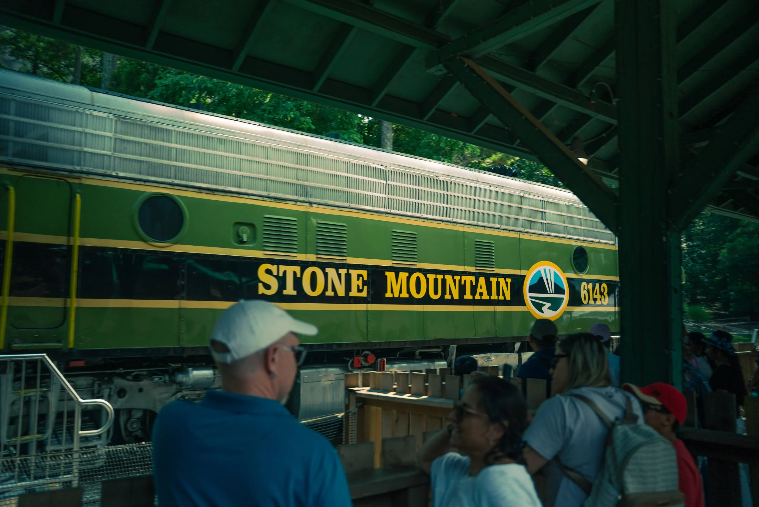 People standing underneath a green train with 'Stone Mountain' written on the side, at Stone Mountain Park, Georgia.