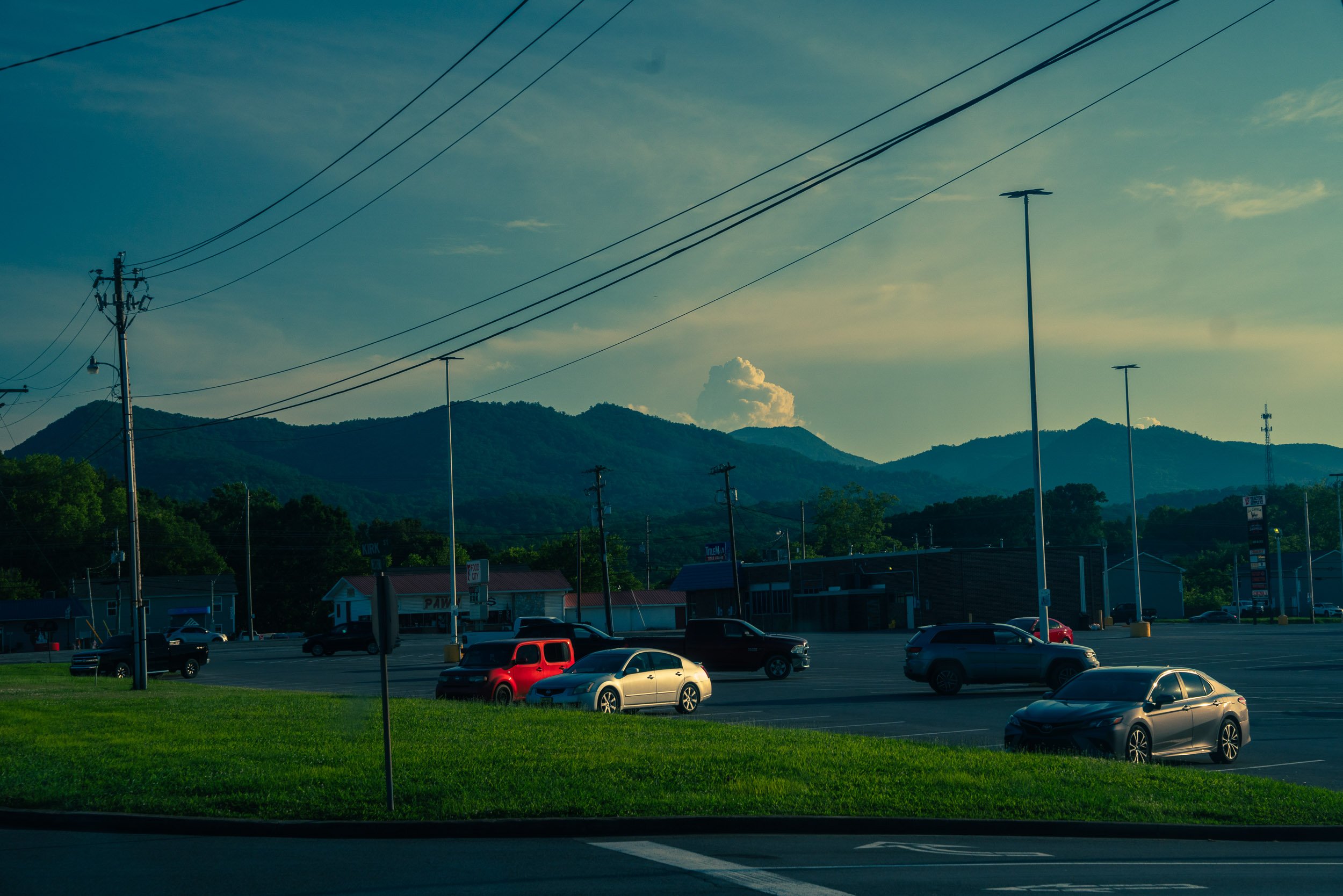Parking lot with cars, surrounded by small buildings, utility poles, and power lines. Hills and clouds visible in the background under a partly cloudy sky.