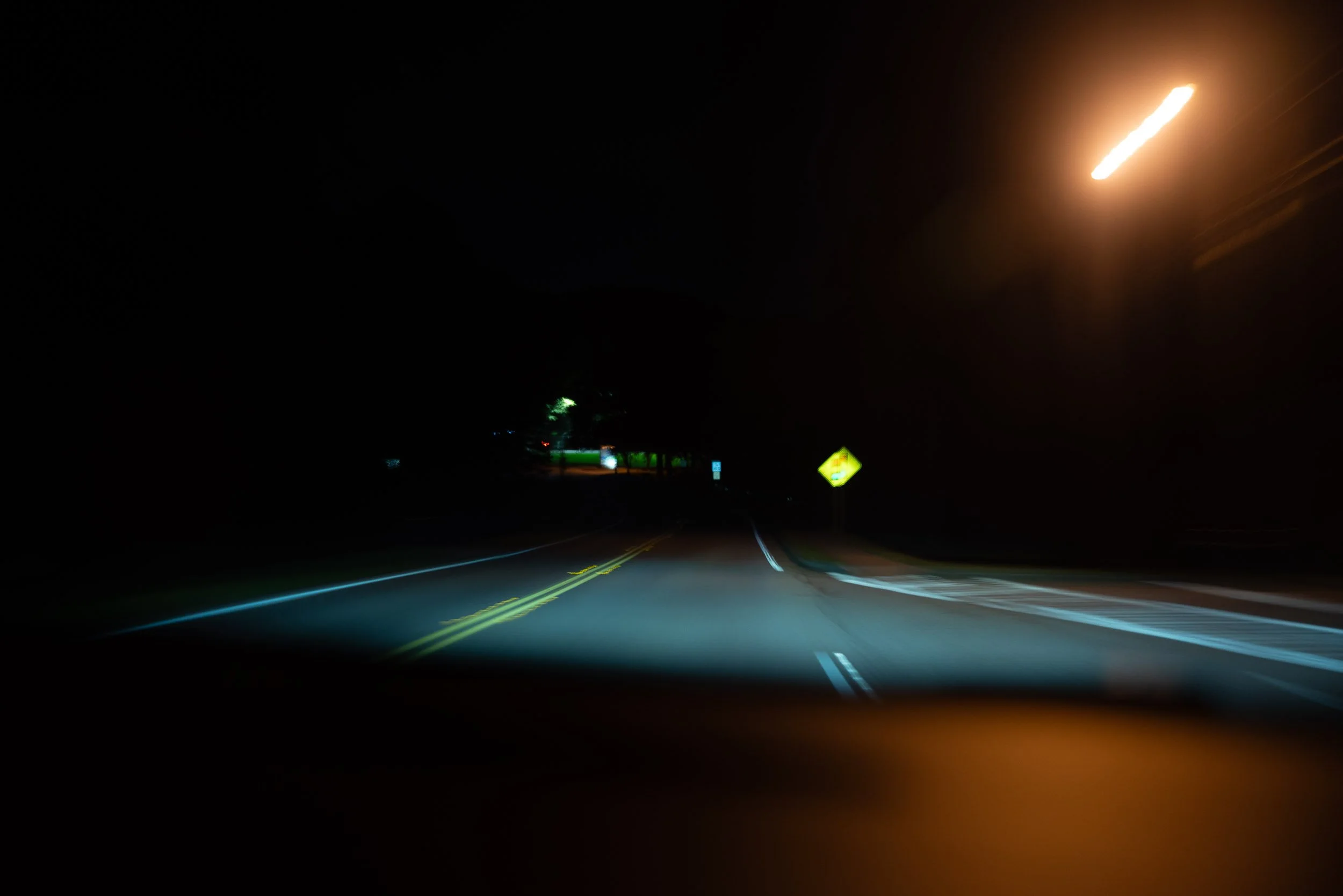 Nighttime view of a dark road illuminated by car headlights with yellow and white road markings, and a green roadside sign in the distance under a dark sky, with blurred light streaks from moving vehicles.