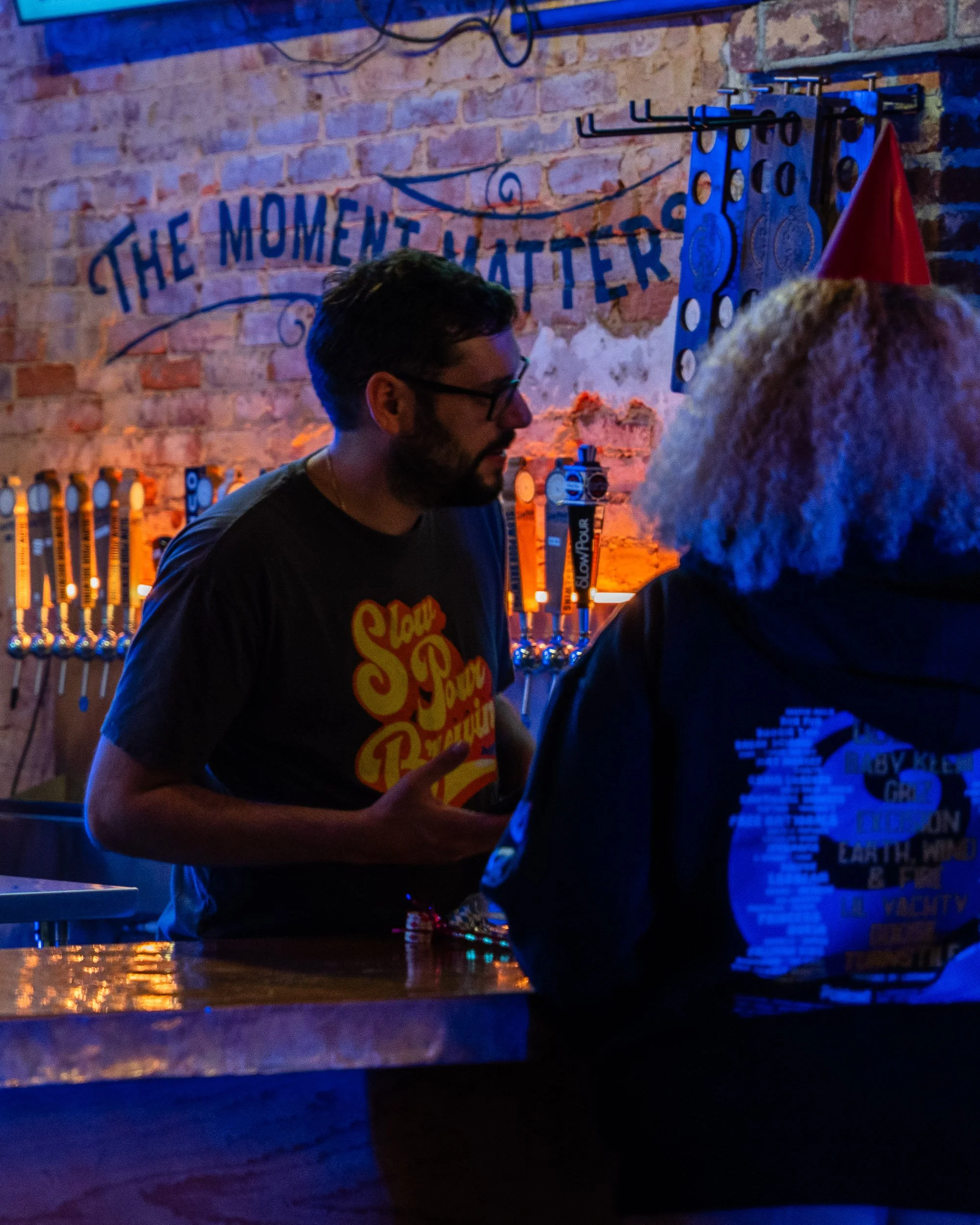 A man with glasses and a beard is standing at a bar, talking to someone whose head is partially visible. The bar has a neon sign that reads 'The Moment Matters,' brick walls, and various hooks and supplies hanging on the wall.