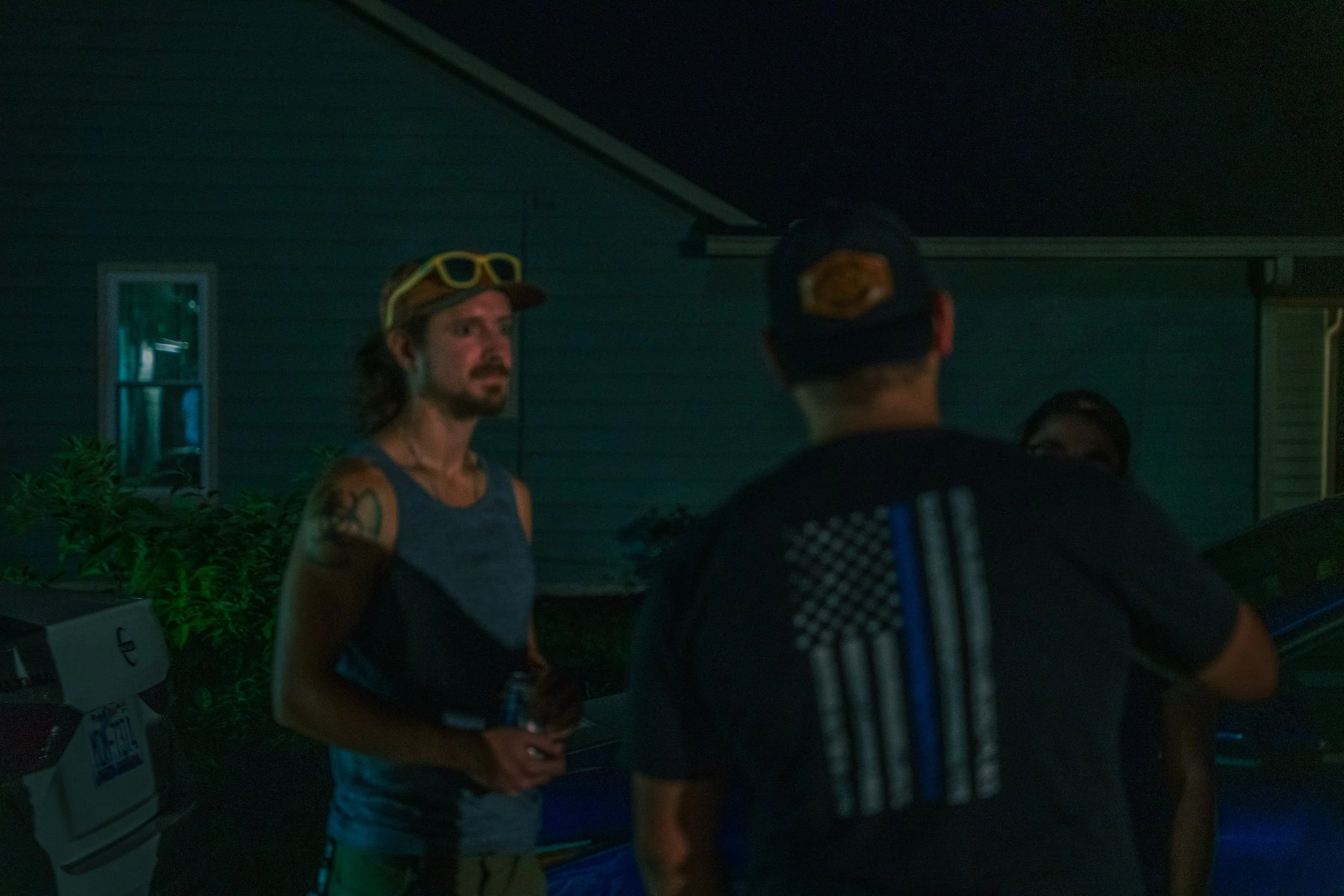 Two people conversing outside near a house at night, one wearing a cap and goggles, the other wearing a cap and a sports shirt, with a window and plants in the background.