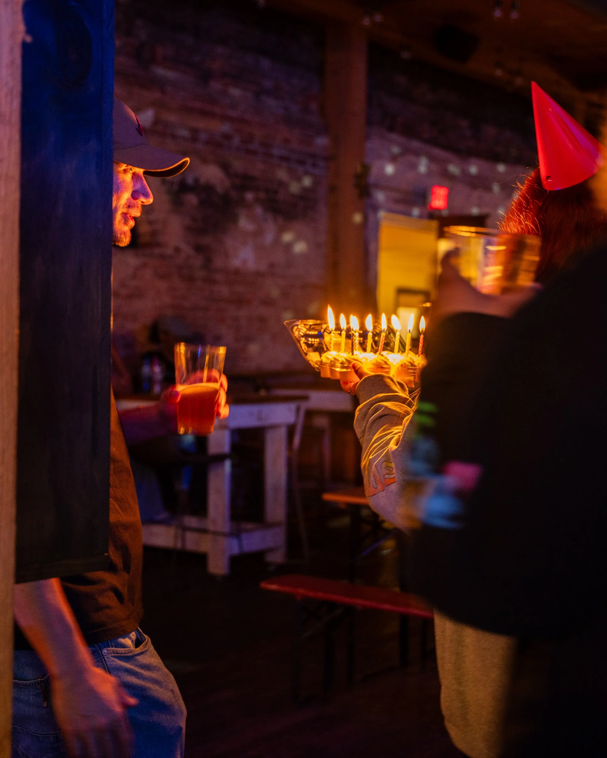 A man in a baseball cap and casual clothing is seen in profile, standing near a wall, with somebody else partially visible on the right side of the image. The person on the right is holding a tray with lit birthday candles, and there is a person wear