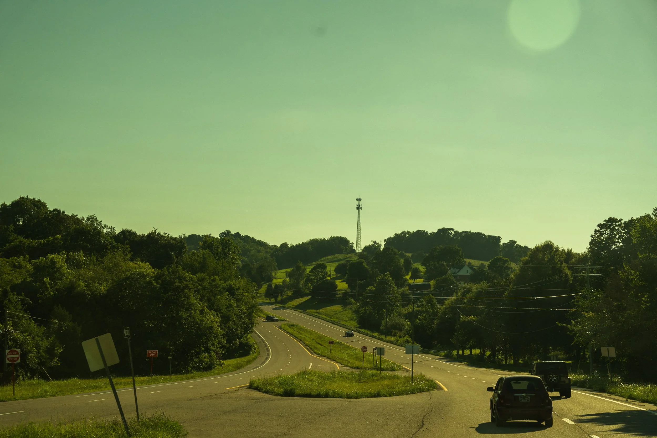 A winding road through a lush green landscape with trees on both sides, with a few vehicles traveling on it and a communication tower on a hill in the distance under a clear sky.