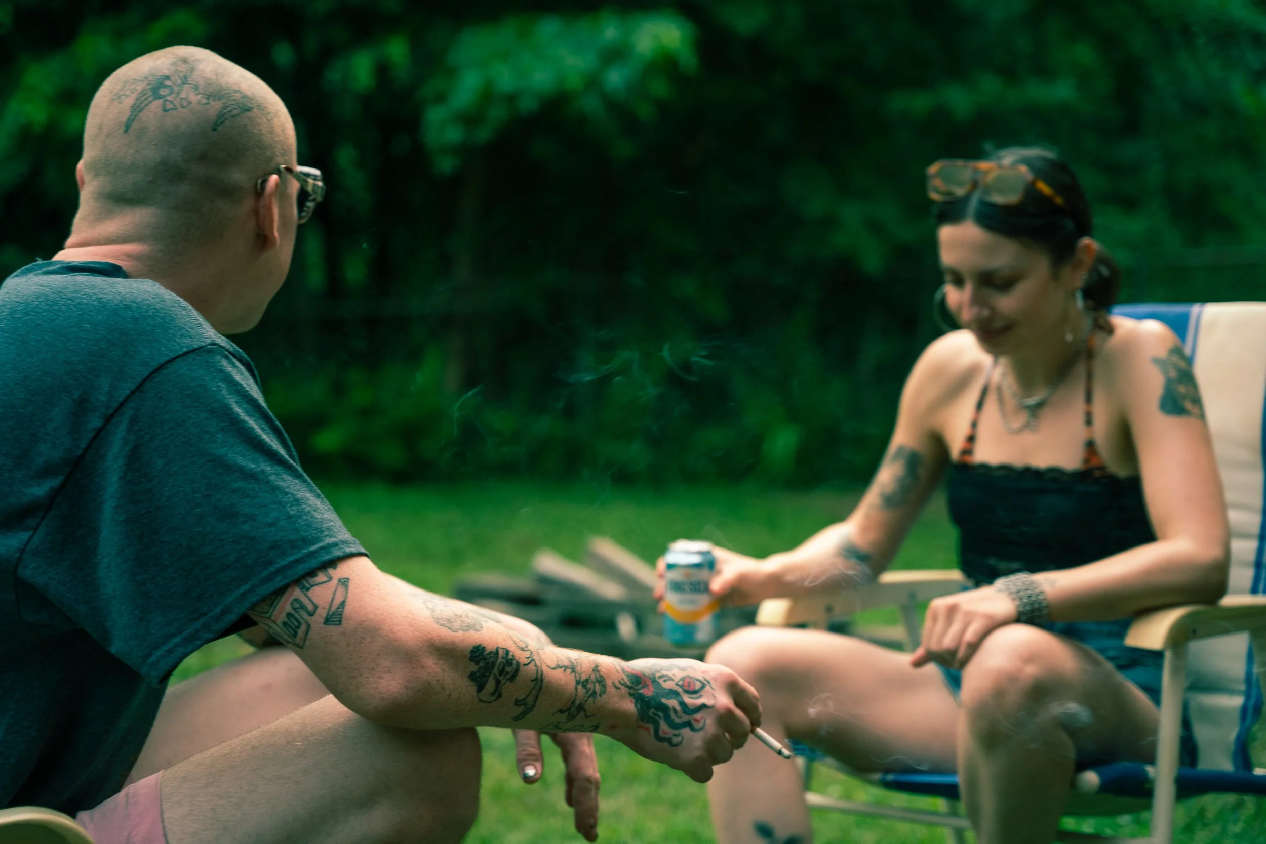 Two tattooed individuals sitting outdoors at a table, engaging in a conversation or activity on a sunny day, with lush green trees in the background.