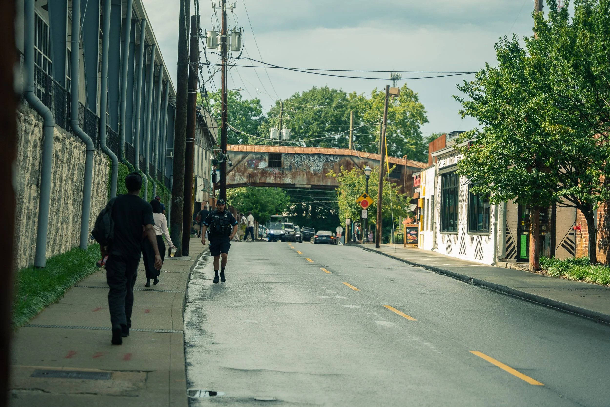 A city street with pedestrians walking on the sidewalk and a few cars parked along the side. There are trees lining the sidewalk and old buildings, including a rusted bridge structure overhead and a building with large windows. Utility poles and wire