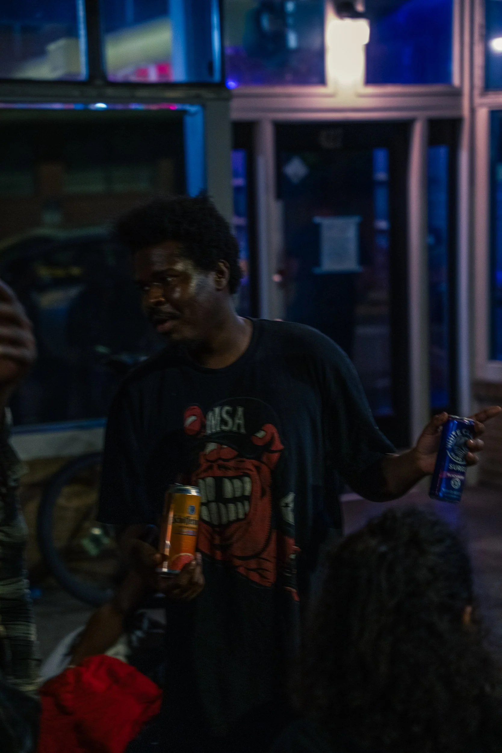 A group of people standing outside at night, with one person holding a bag of snacks and a drink. The background features a building with large glass windows and blue lighting.