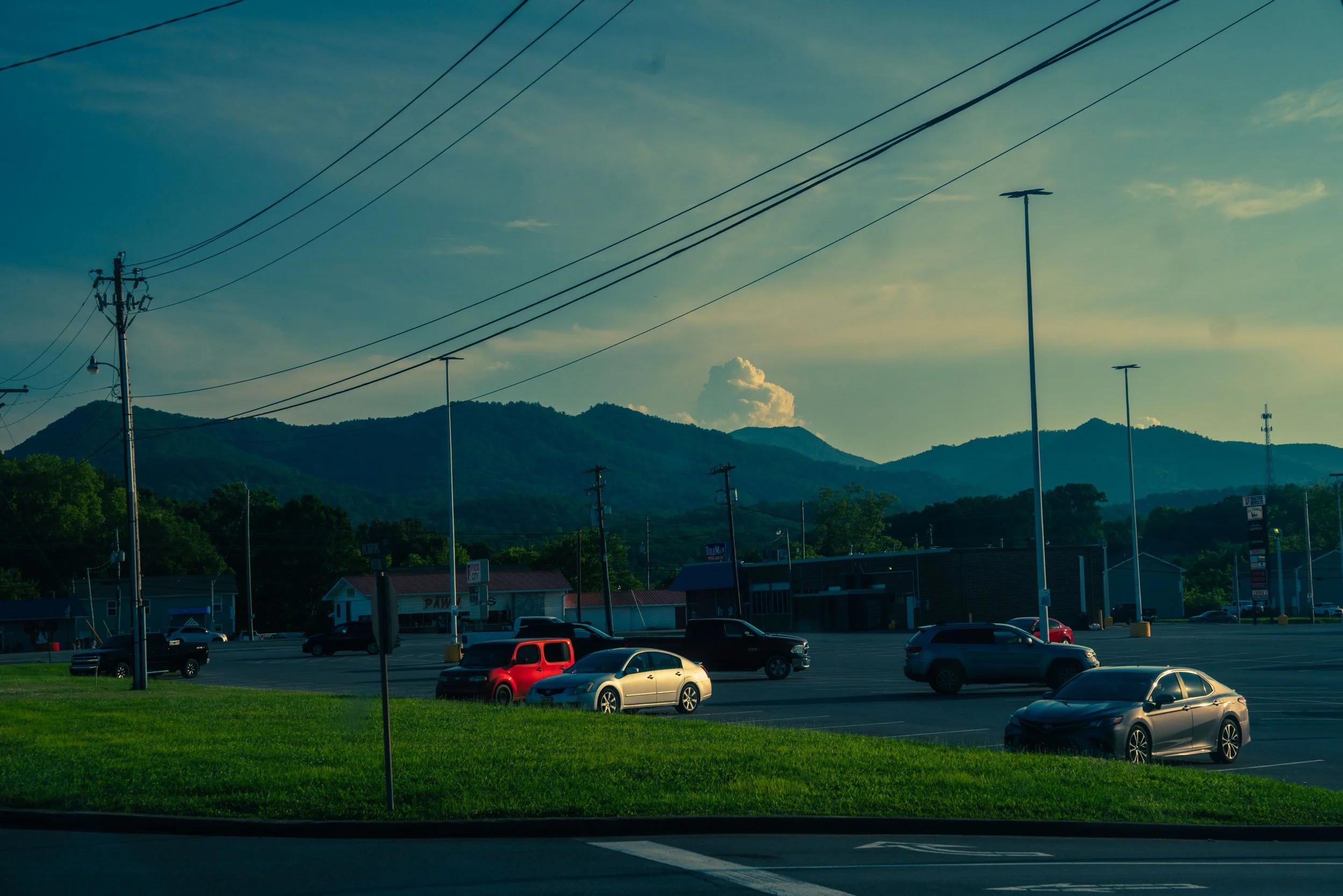 A parking lot with several parked cars, commercial buildings, utility poles with power lines, a mountain range in the background, and a cloudy sky.