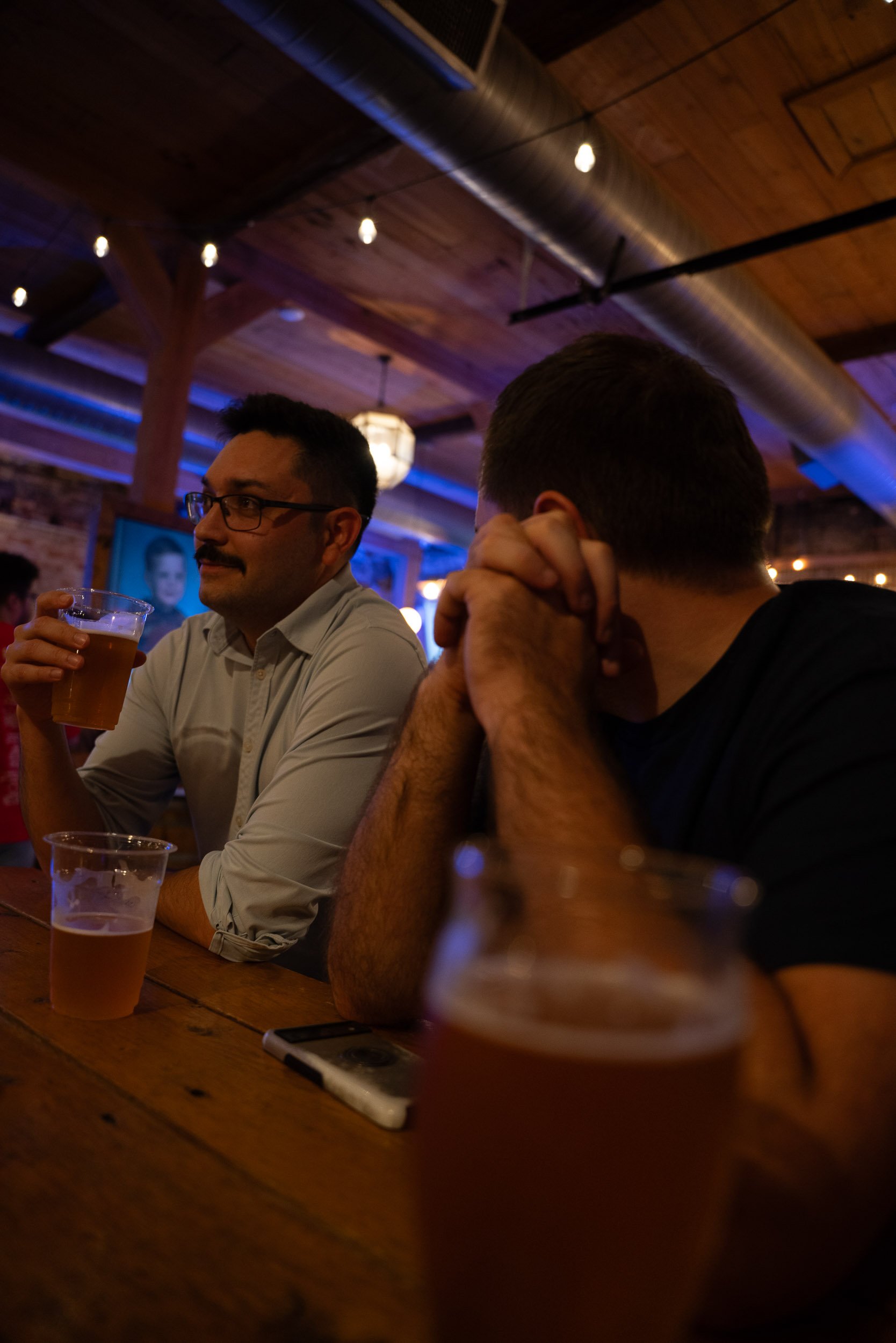 Two men sitting at a wooden table in a dimly lit bar or restaurant, one holding a pint of beer and the other resting his head on his hand, with a blurred background of exposed brick walls and hanging lights.