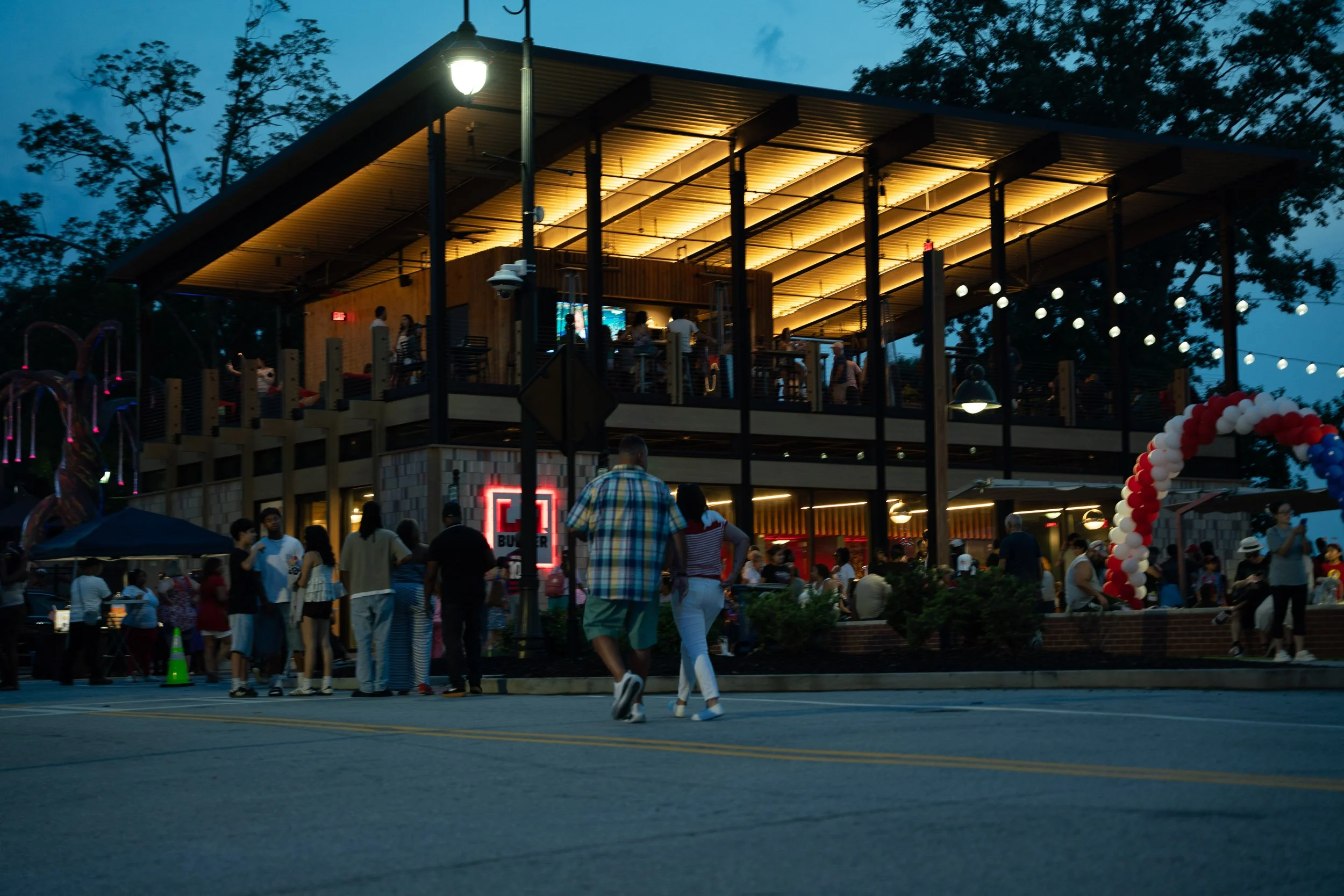 A crowd of people gathered outside a two-story building at dusk. The building has a modern design with a large, illuminated roof and people inside. There are trees in the background, and an arch made of red, white, and blue balloons in the lower righ
