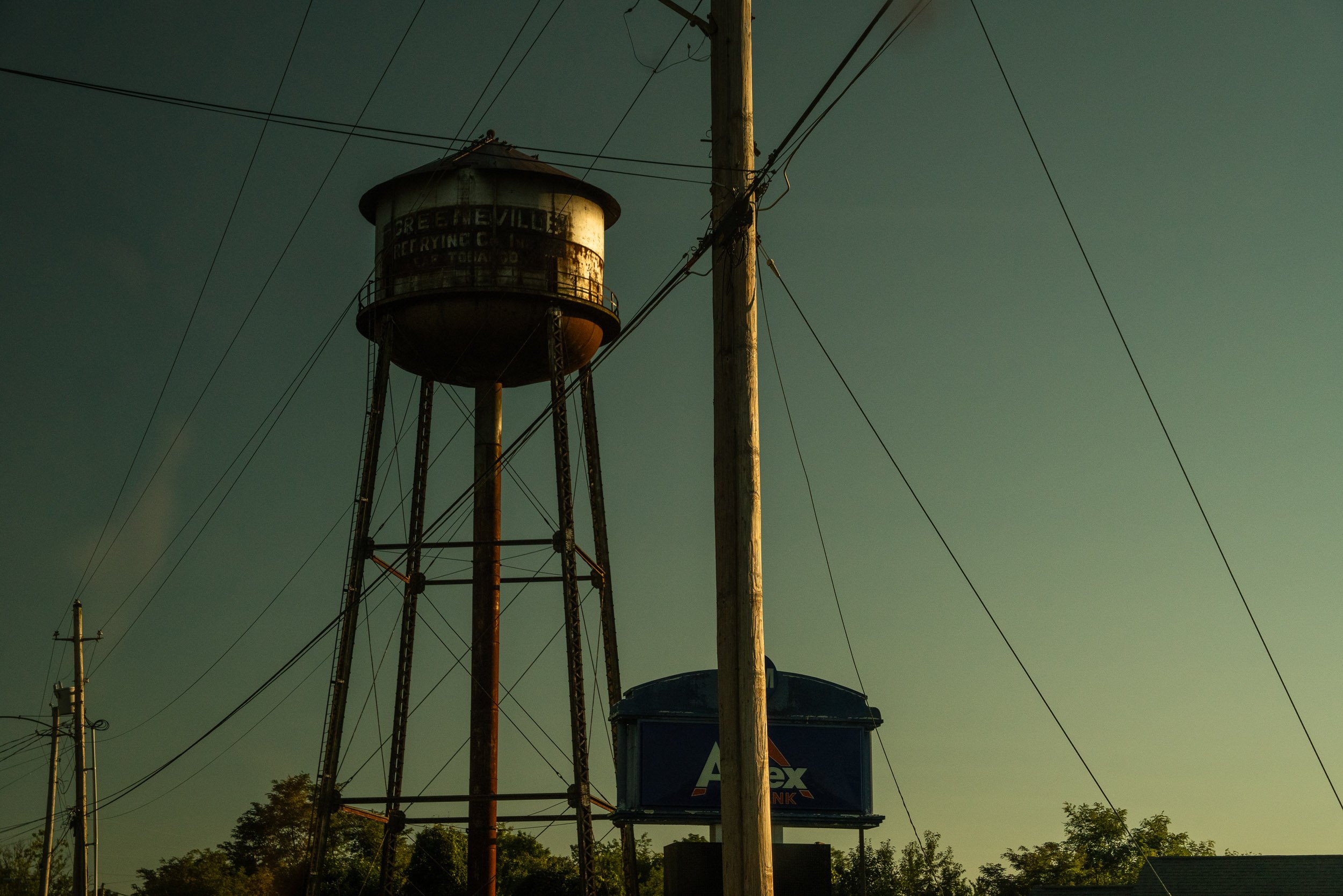An old water tower with a rusted metal exterior, standing tall among utility poles and wires against a cloudy sky, with trees in the background.