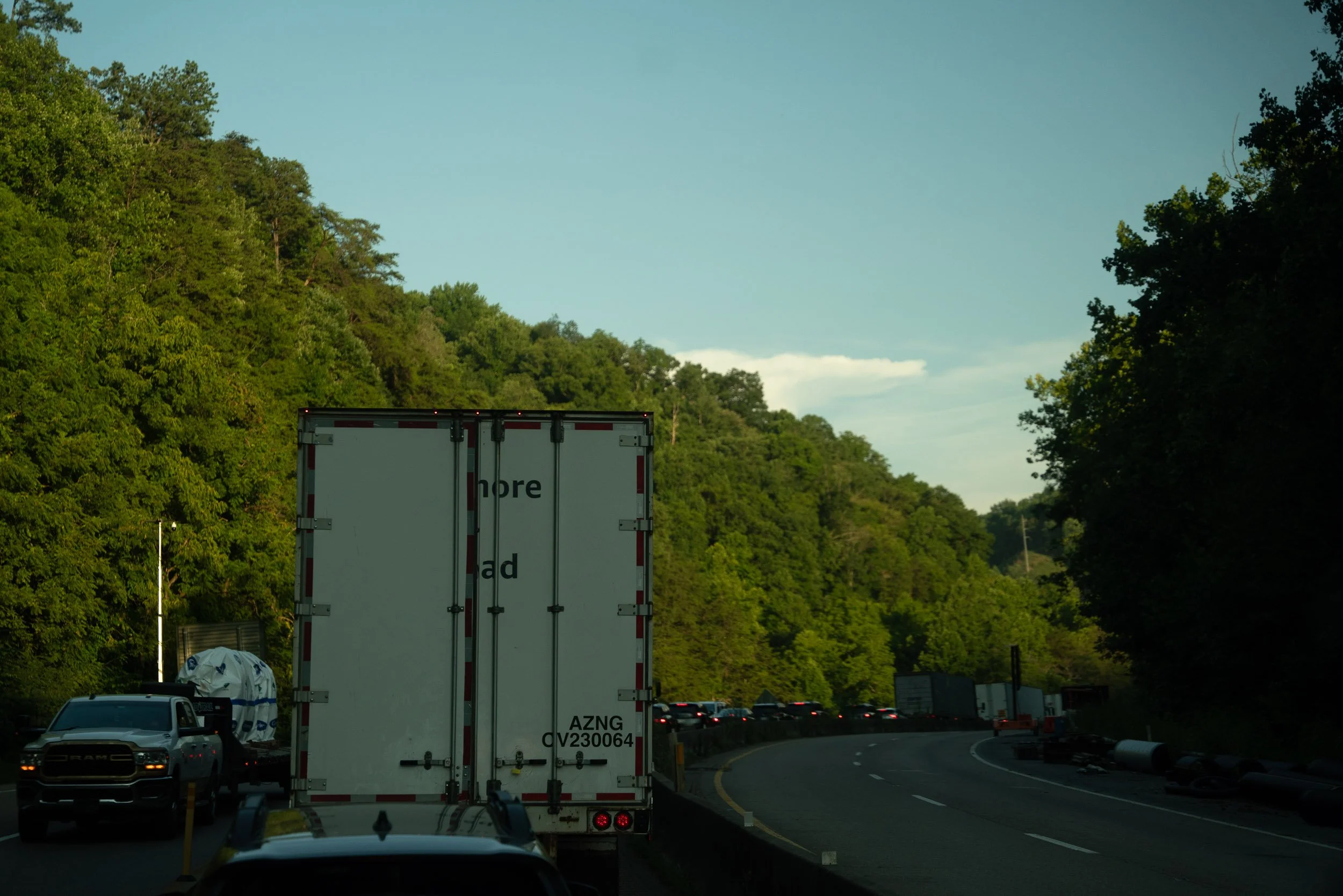 Traffic jam on a highway with trucks and cars, surrounded by green trees and a clear blue sky.