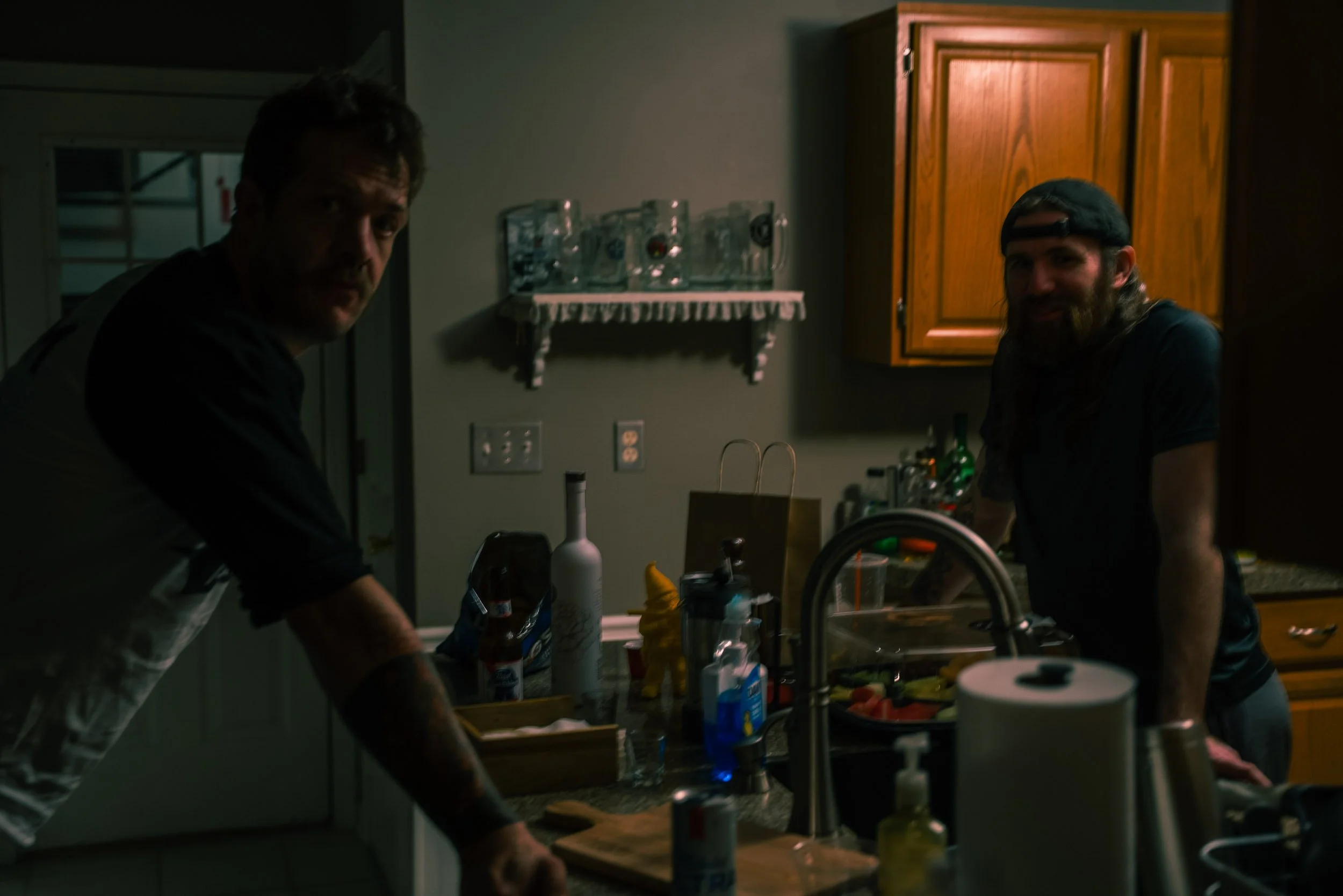 Two men are in a kitchen, one on the left wearing a white apron and the other on the right with a dark shirt and backwards cap, surrounded by various kitchen items on the countertop.