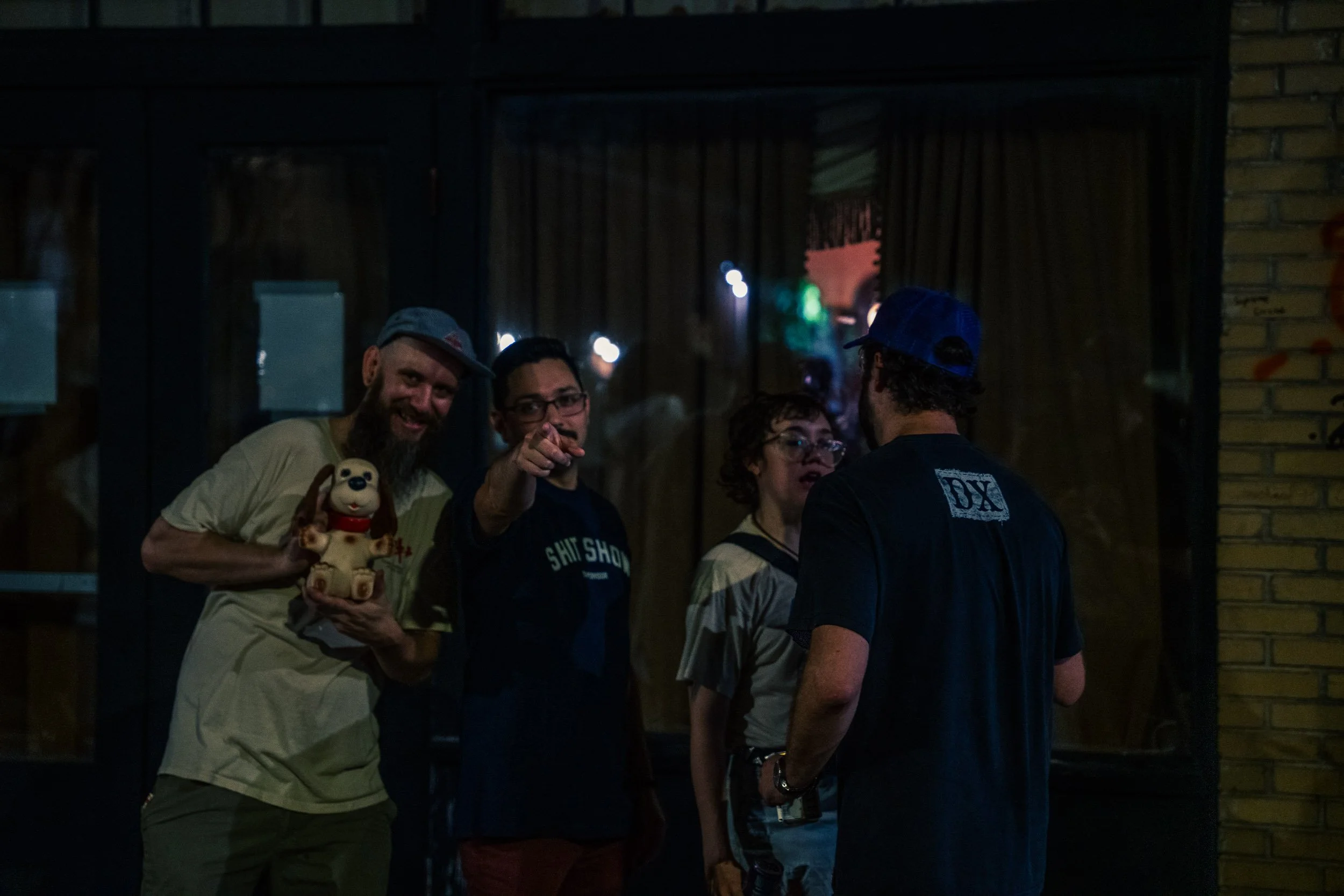Four people are standing and talking outside in the evening. One man in a gray cap and beige shirt is holding a small plush toy. They are near a wooden wall and a brick wall.